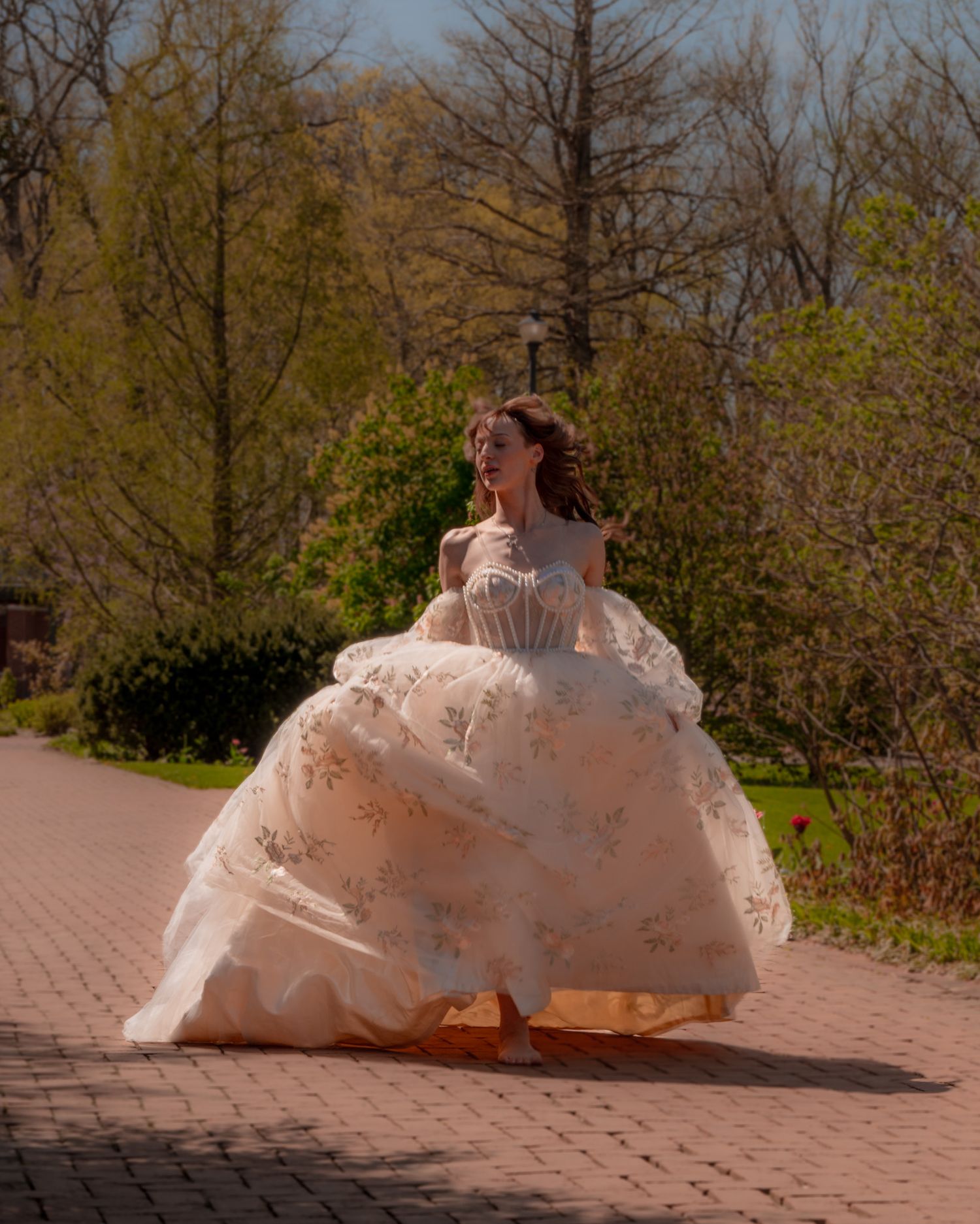 A bride twirls in a voluminous floral ball gown on a brick path surrounded by lush spring trees.