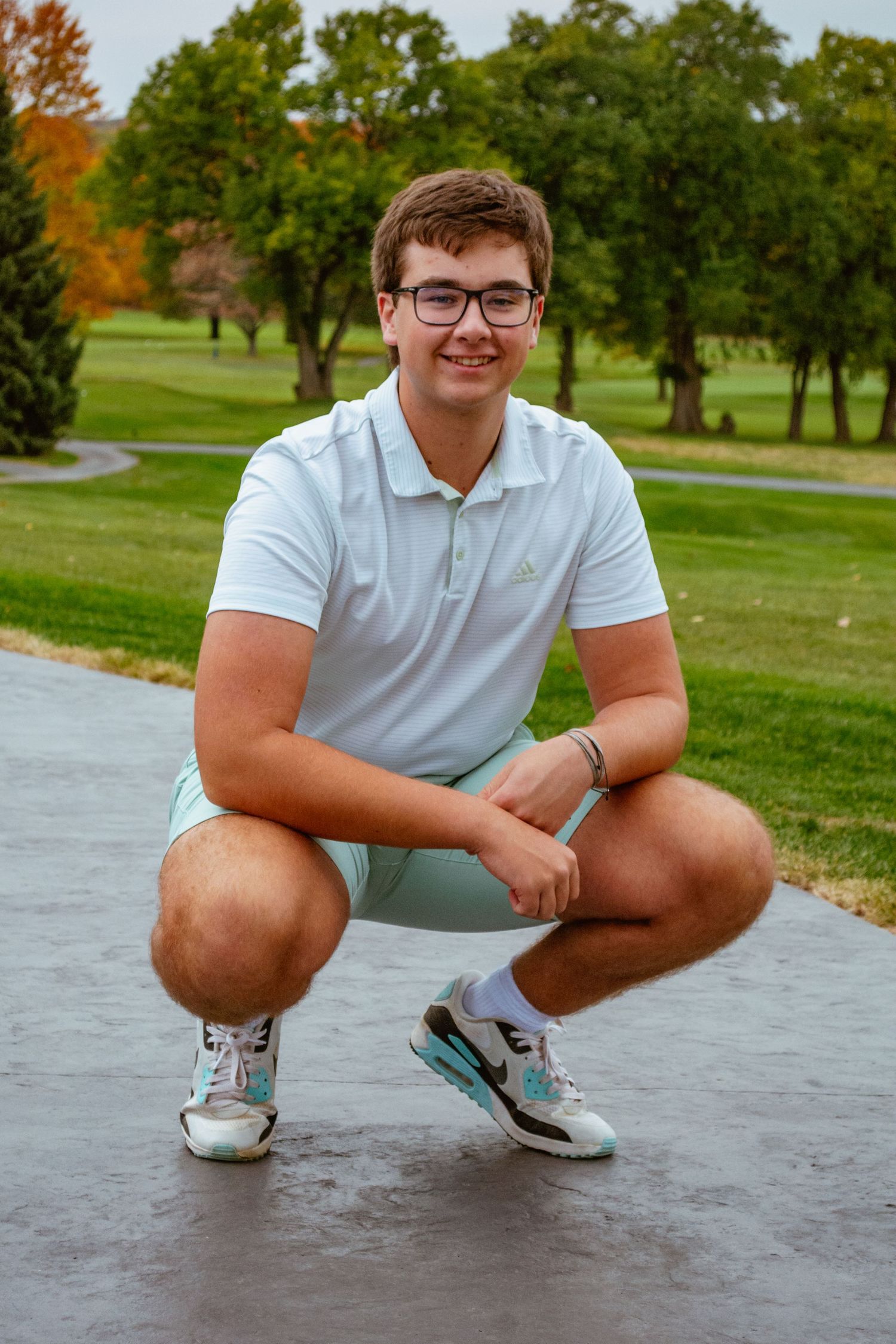 A person in a light blue polo shirt and shorts crouches on a path with autumn trees in the background.