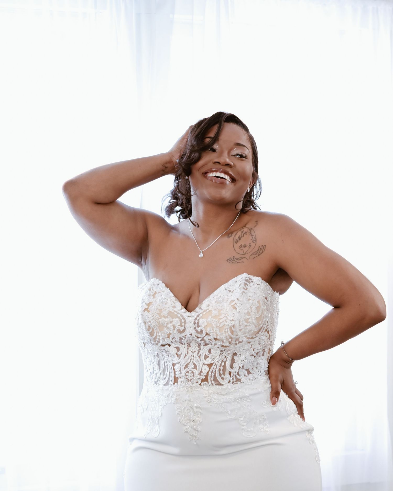 Smiling bride in a white lace strapless wedding gown posing confidently near a bright window.
