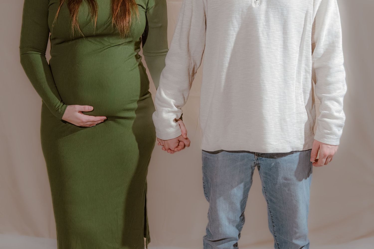 Pregnant woman in green dress holding hands with man in white shirt and jeans during maternity photoshoot.