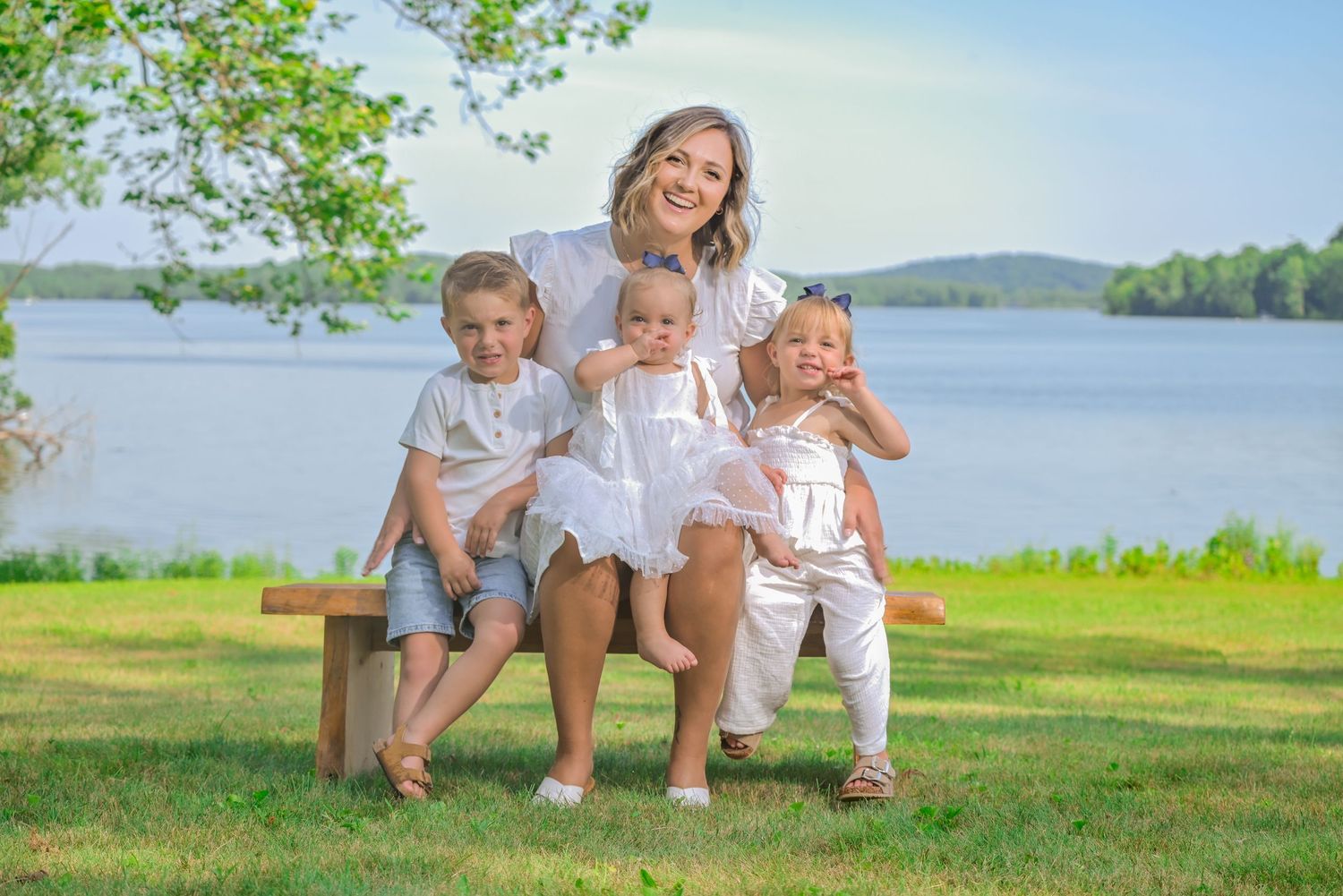 A smiling woman poses on a wooden bench outdoors with three young children near a serene lake on a sunny day.