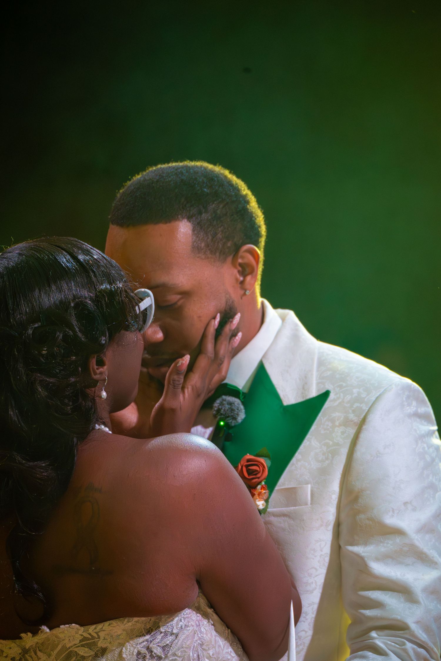 A couple shares a romantic kiss at their wedding, groom in white tuxedo with green accents and orange boutonniere.