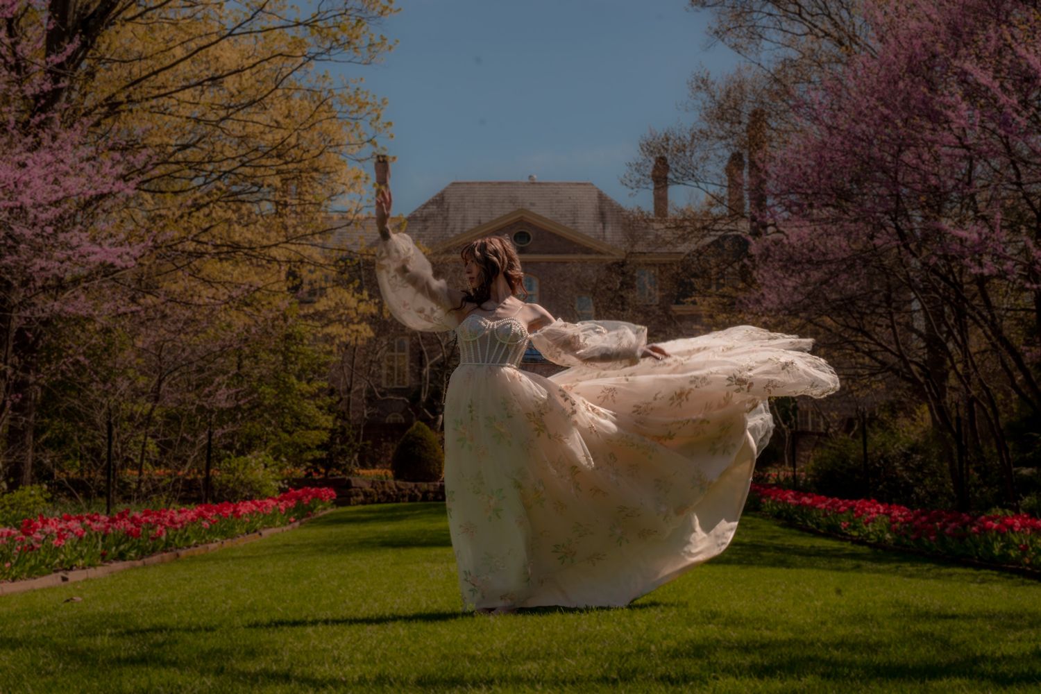 Woman in flowing white gown twirling in a spring garden with blooming trees and tulips near a grand estate.