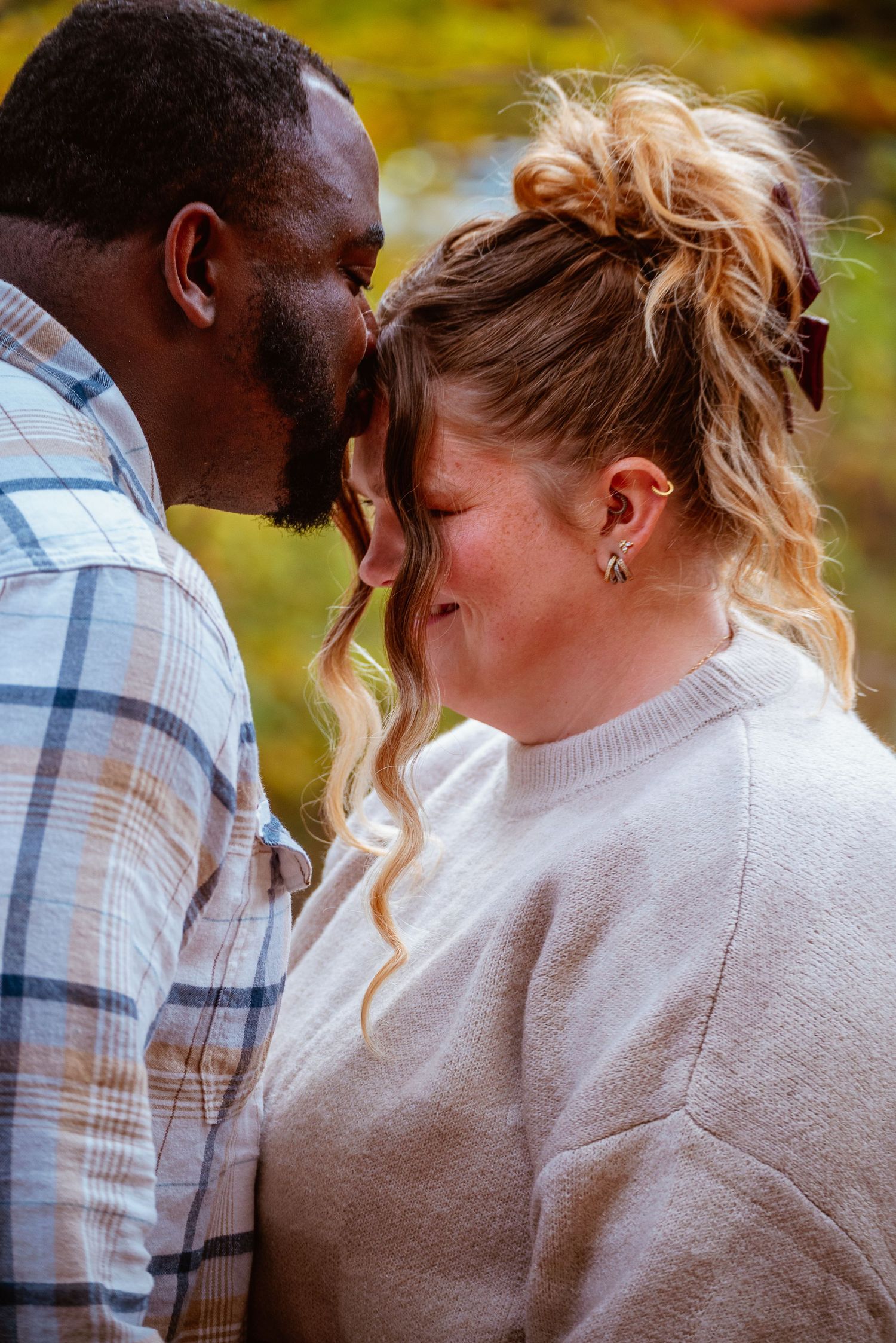 An interracial couple shares a tender forehead-to-forehead moment outdoors in autumn, smiling softly.