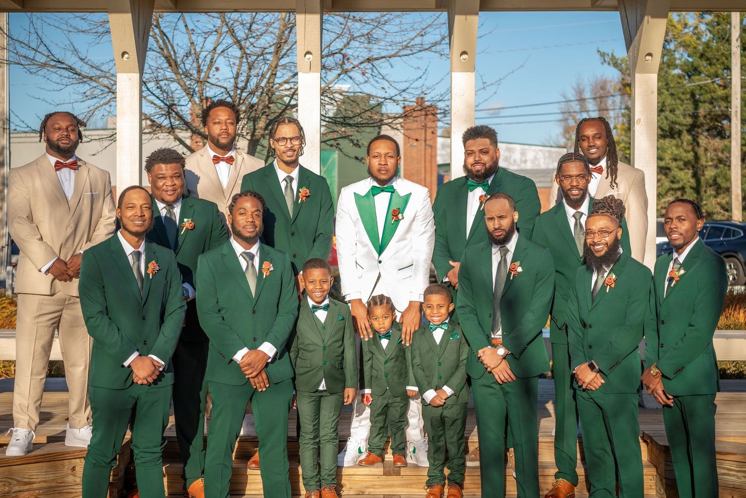 Groom in white tuxedo poses with groomsmen in green suits and two young boys at an outdoor wedding ceremony.