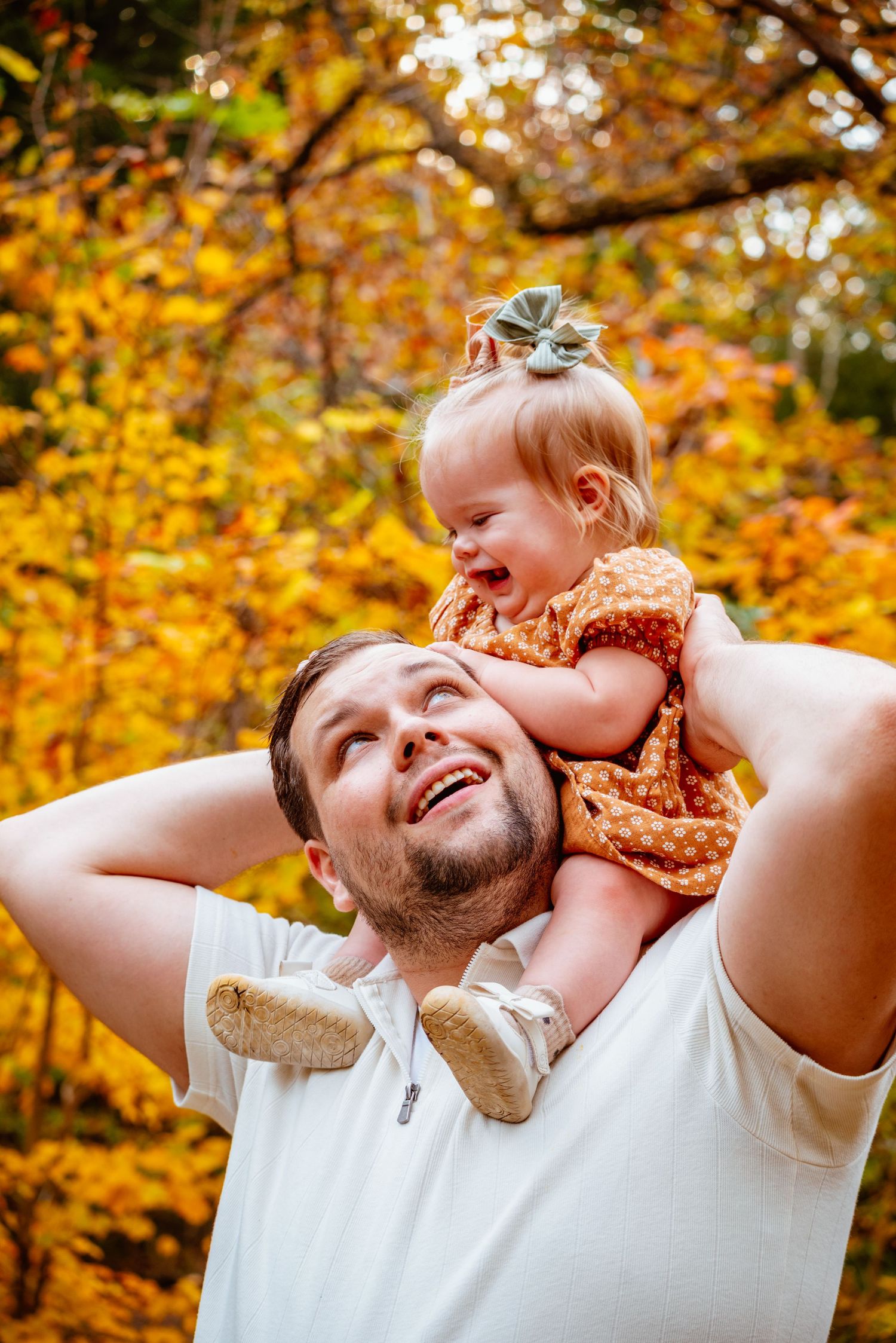 A smiling father holds his laughing toddler girl on his shoulders amid vibrant autumn foliage in a fall park.