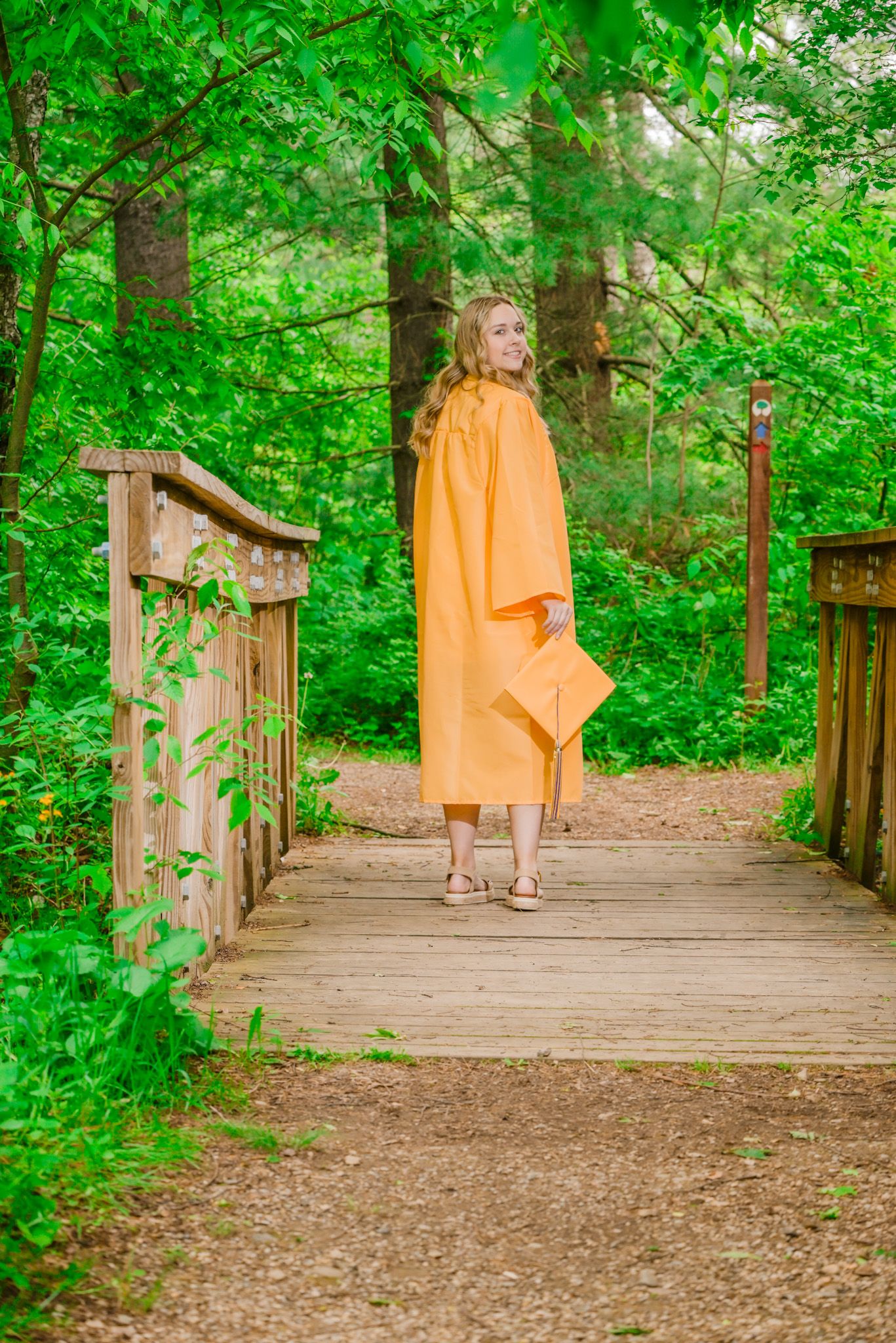 Young woman in orange graduation cap and gown standing on wooden bridge surrounded by lush green forest.