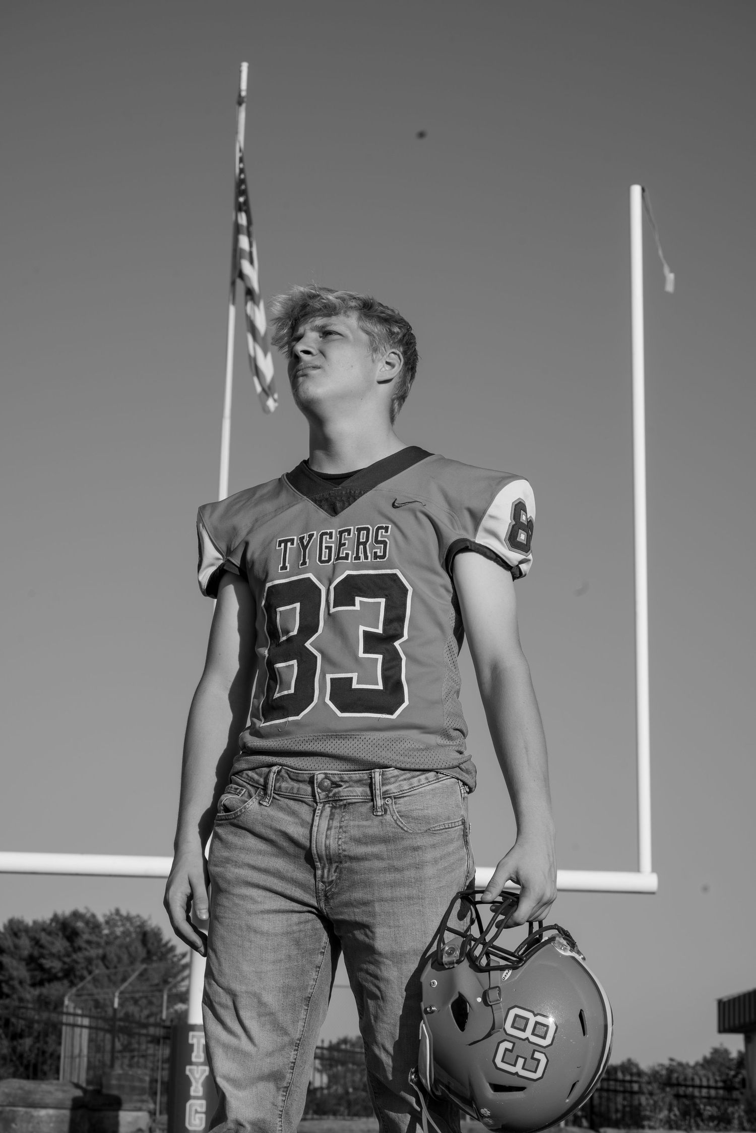 Black and white photo of a teen football player in Tygers #83 jersey holding helmet on field with goalpost and American flag.
