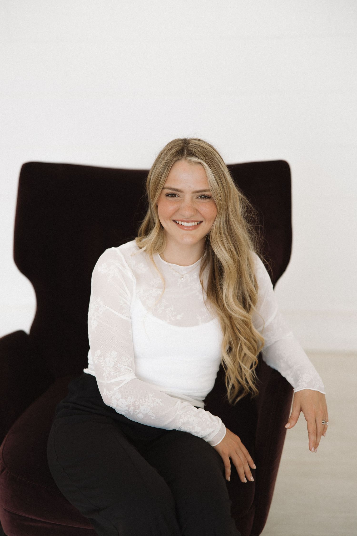 Professional in white blouse seated in dark velvet chair against white background.