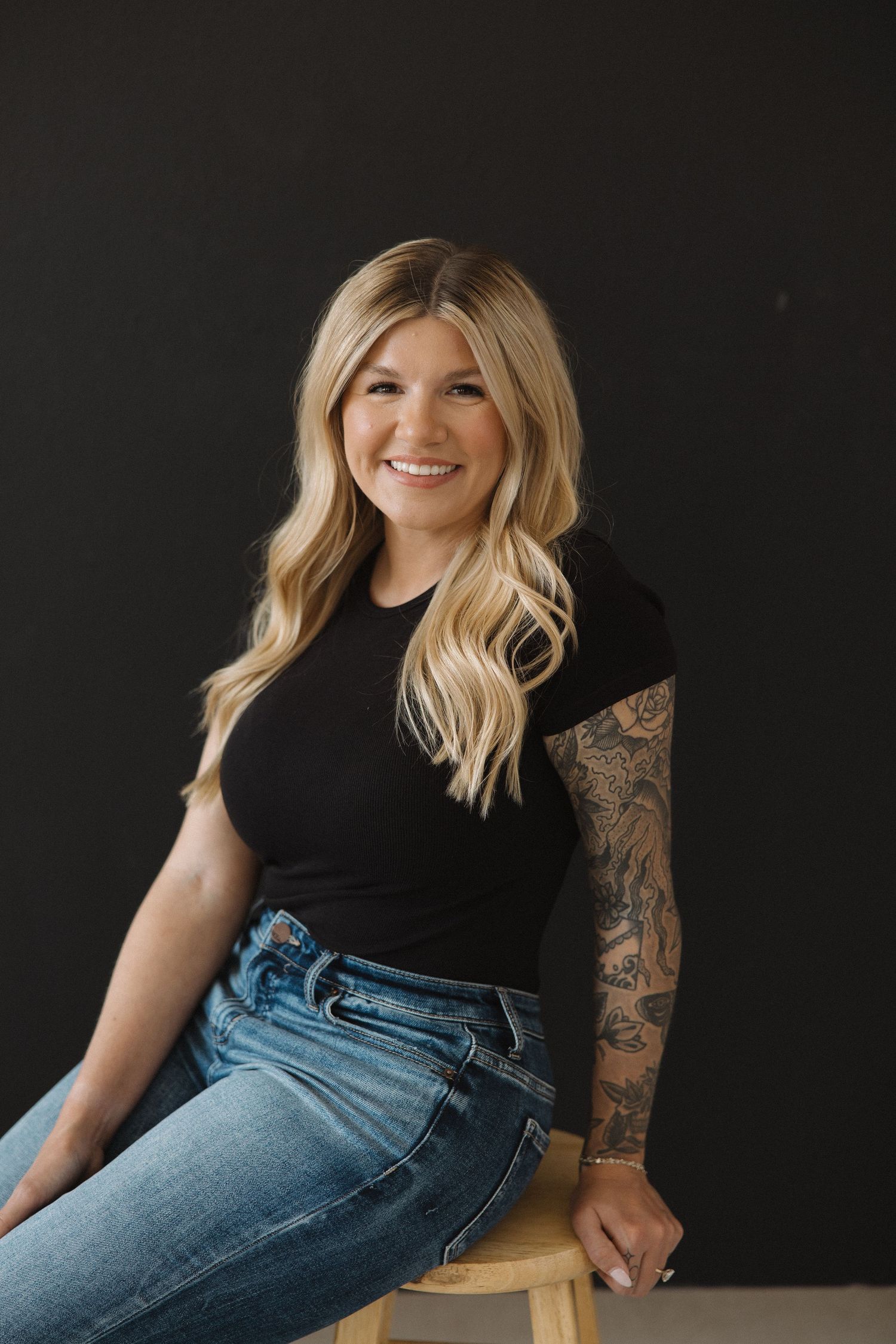 Portrait of someone in black top with jeans and tattoos against dark background.