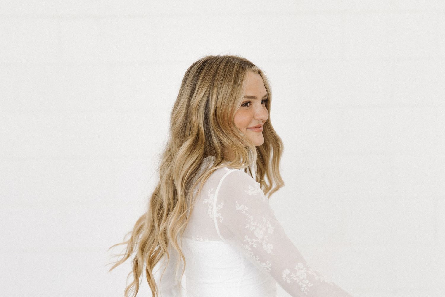 Portrait of person in white lace dress with wavy blonde hair against white background.