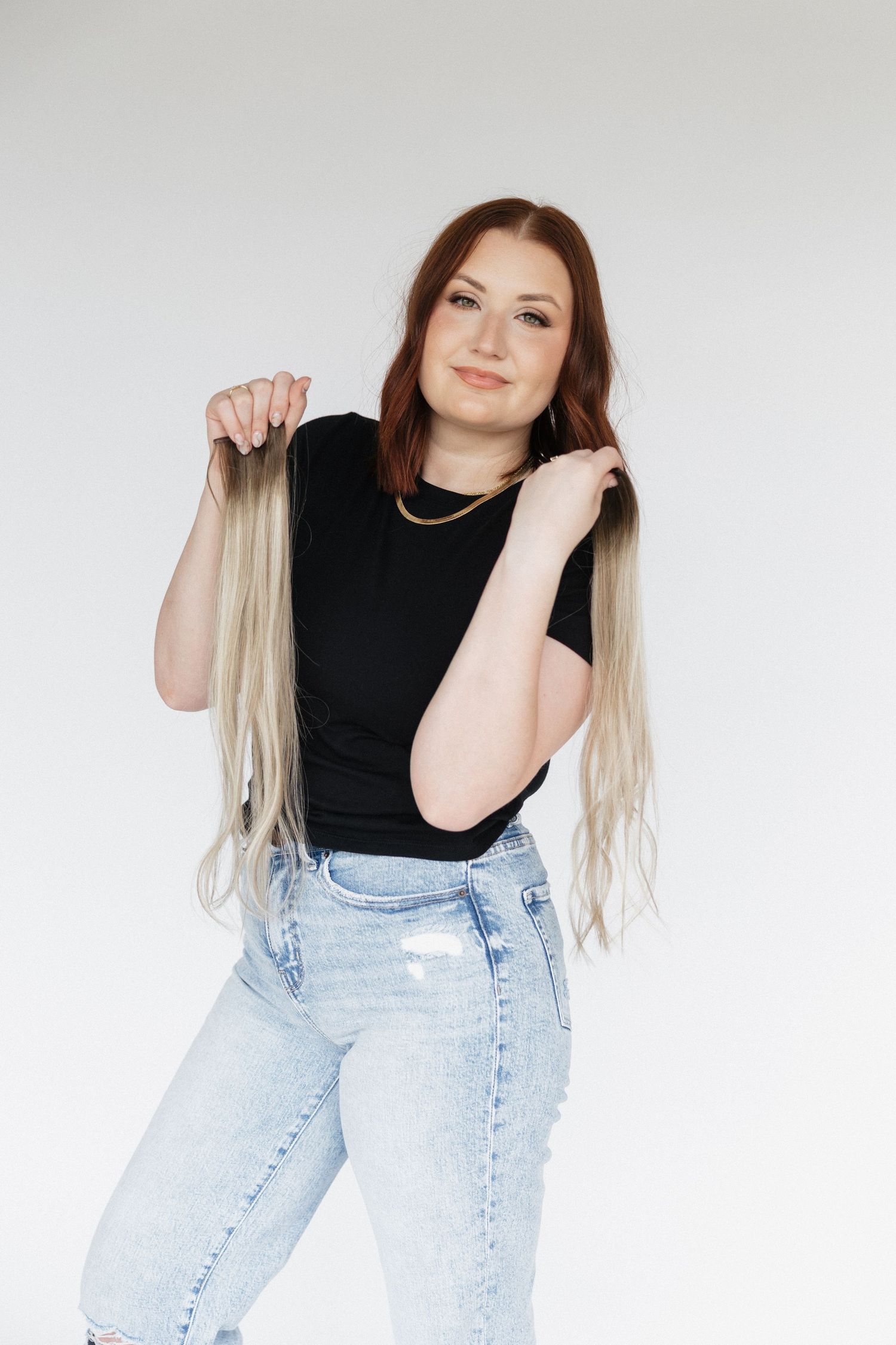 Model in black top and light wash jeans poses casually against white background with hair extensions.