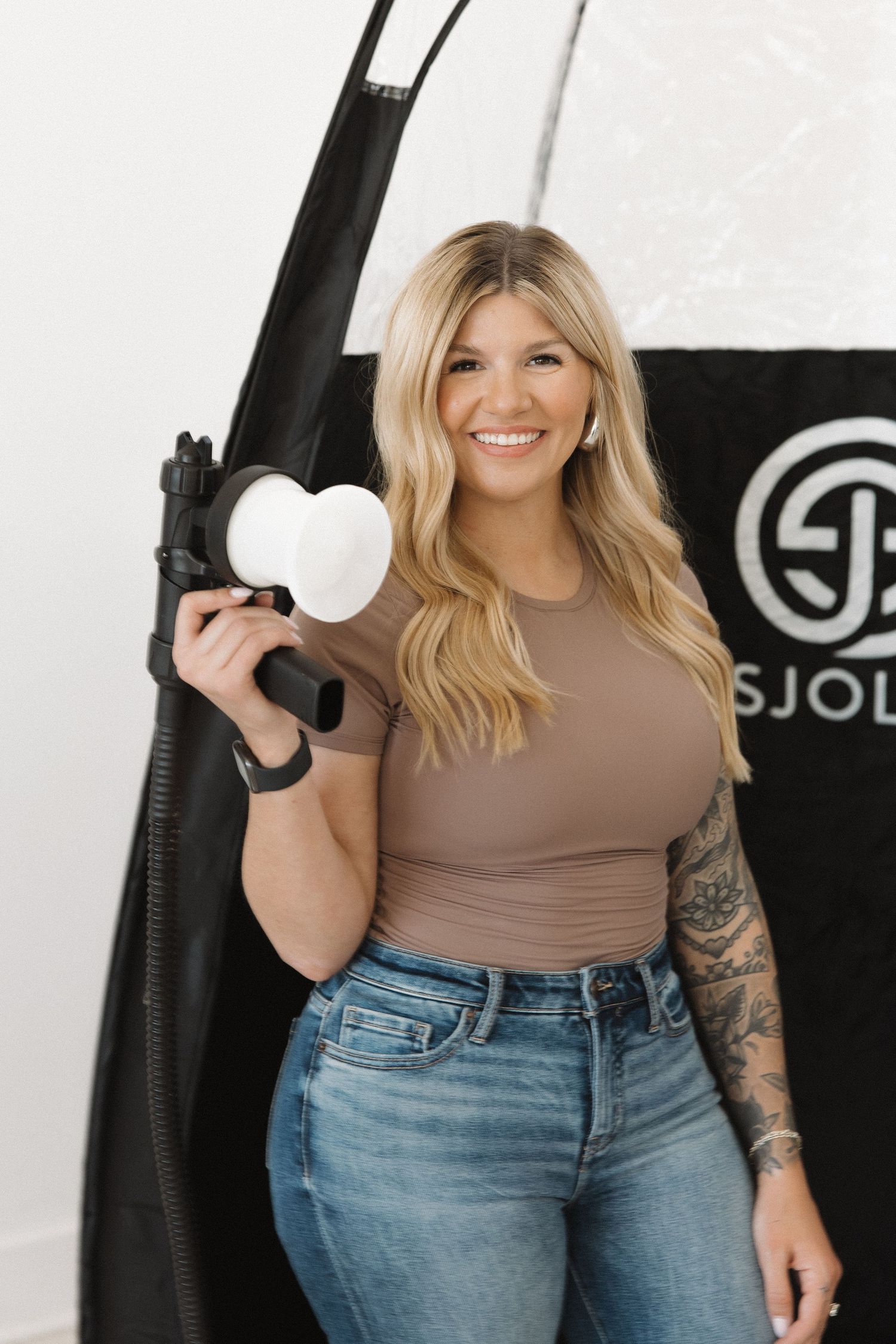 Spray tan professional holding equipment in front of black Sjolie tent backdrop.