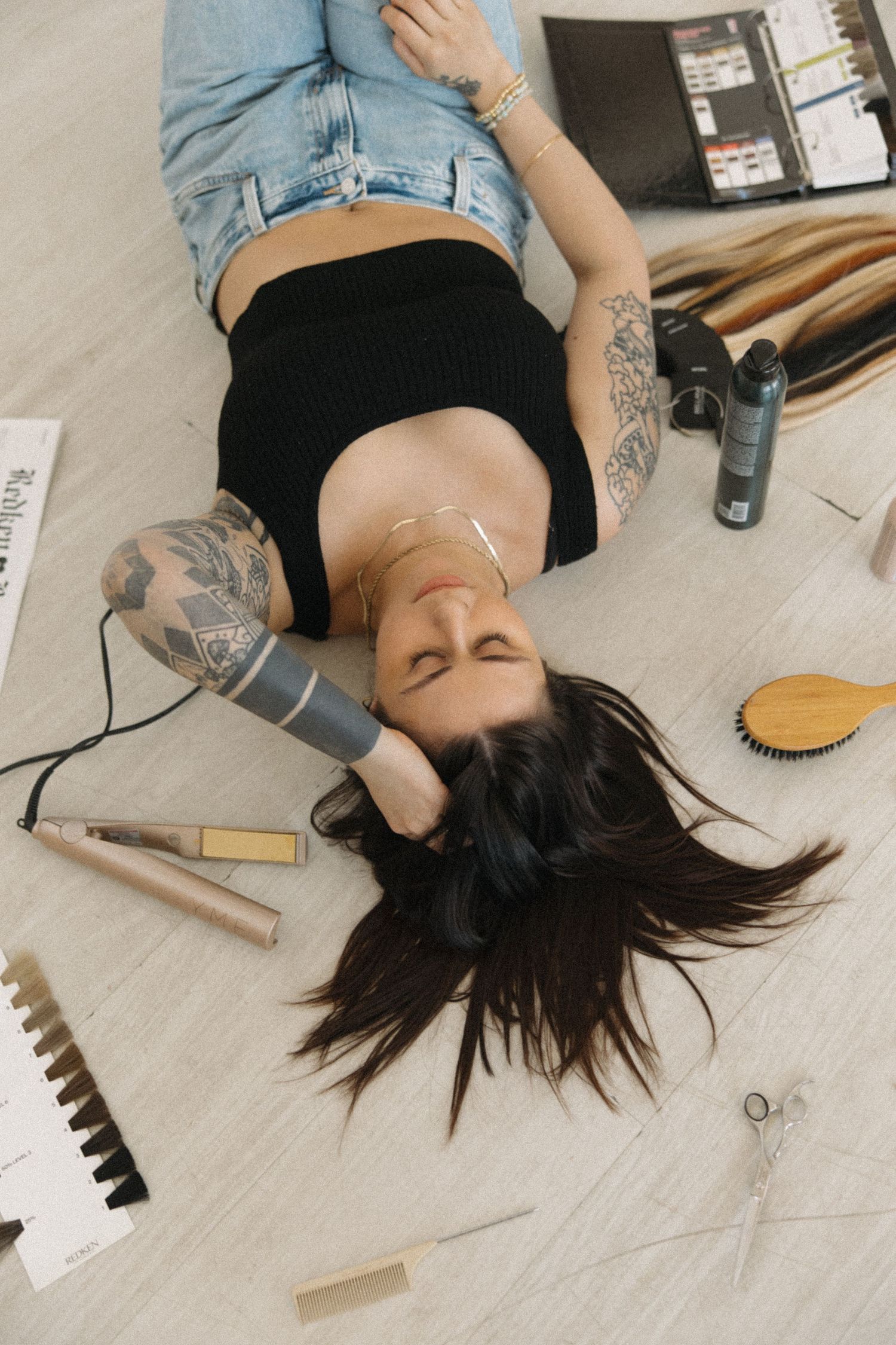 Person lying on floor with beauty products and hair tools scattered around in artistic lifestyle shot.