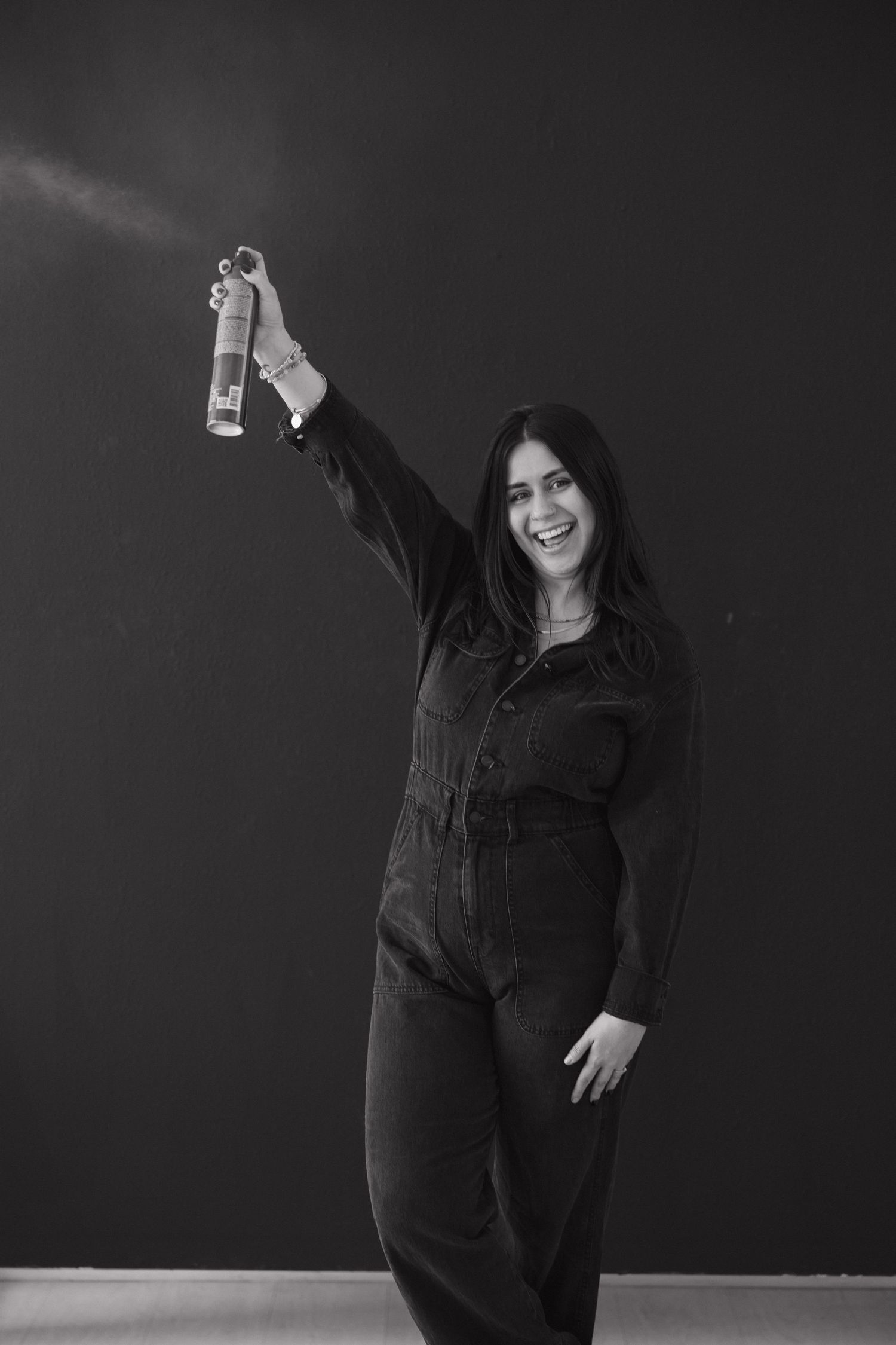 Stylist in black outfit holds up hairspray bottle in black and white photo.