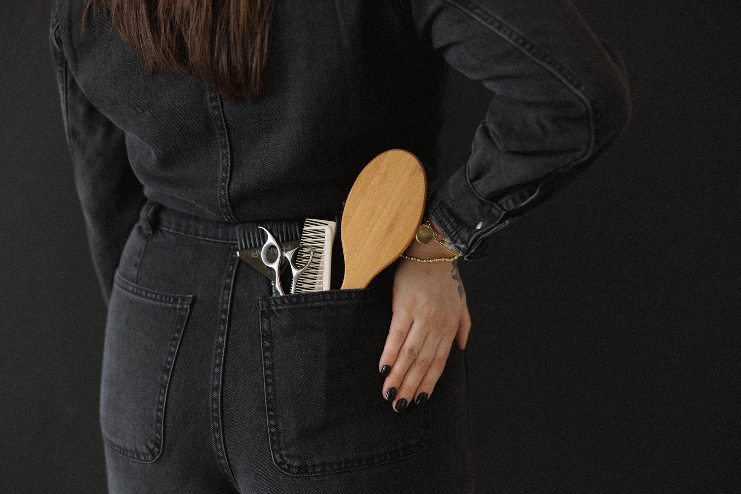 Hair styling tools visible in back pocket of black jeans against dark background.