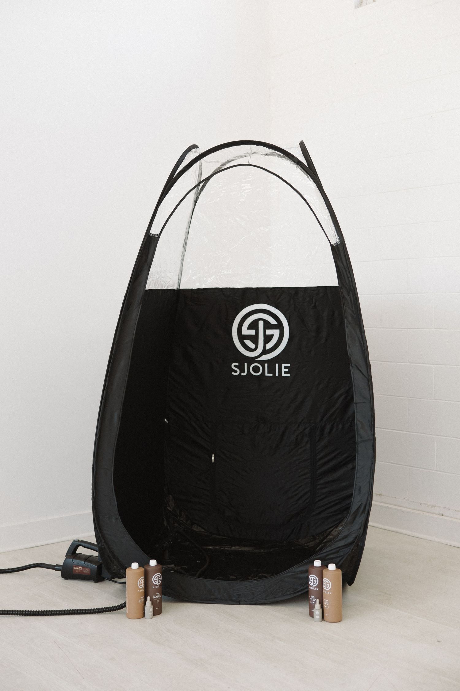 Black pop-up spray tanning tent with SJOLIE logo displayed against white wall.