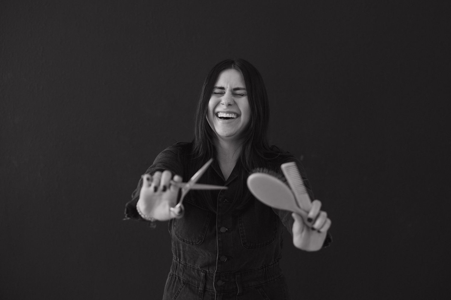 Hairstylist in black outfit playfully poses with styling tools against dark background.