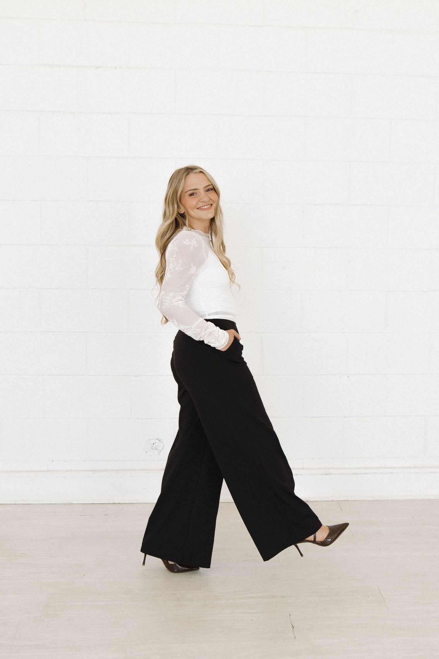 Woman wearing a white blouse and black pants stands against white wall in dynamic pose.