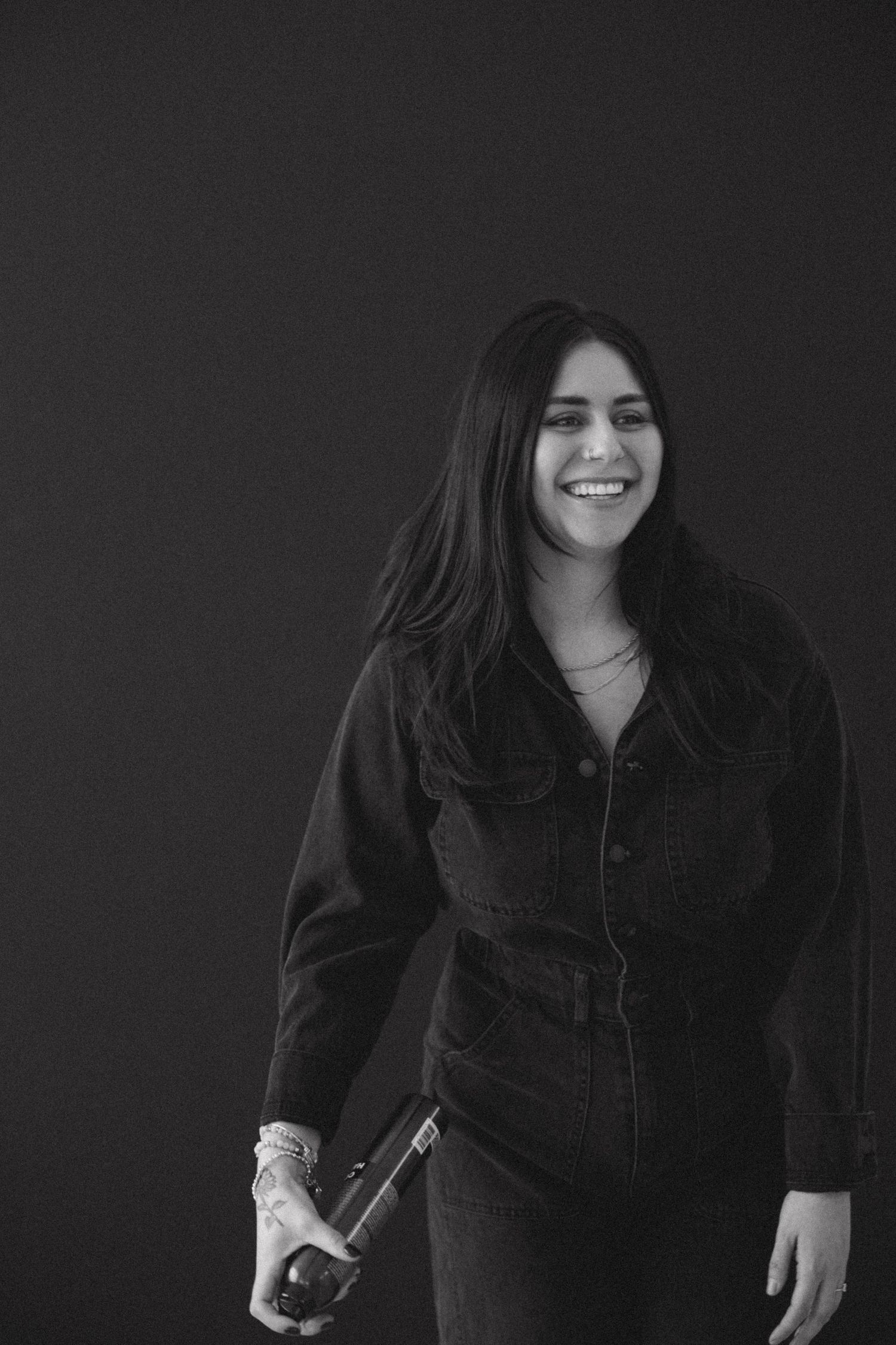 Hair professional in dark jumpsuit holds styling tools in monochrome portrait.