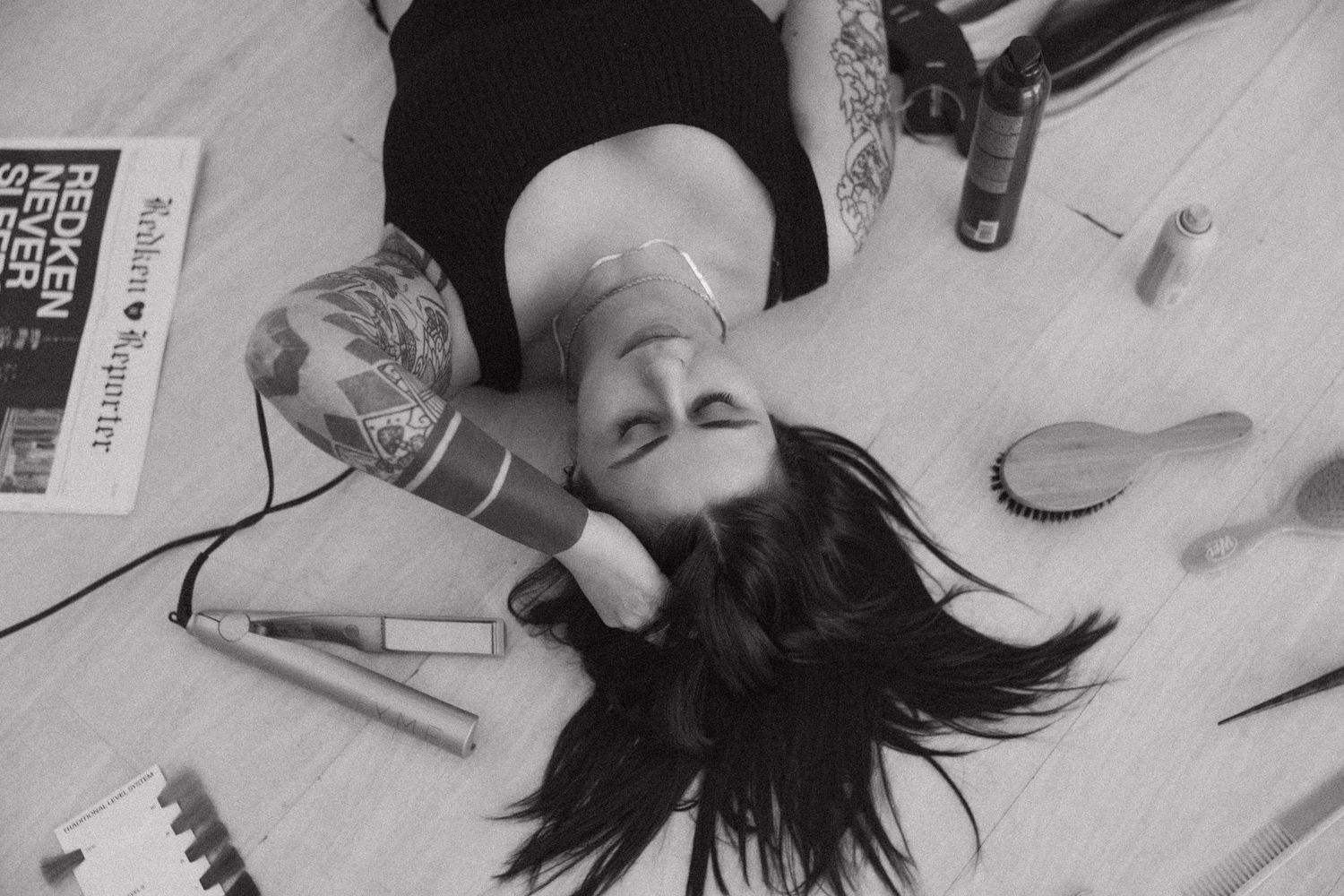 Black and white photo of someone lying on floor among scattered beauty supplies and brushes.