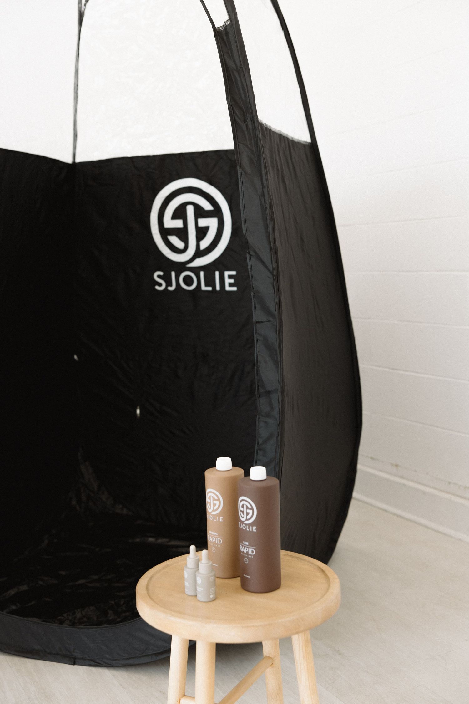 Black Sjolie spray tan tent with product bottles displayed on wooden stool.