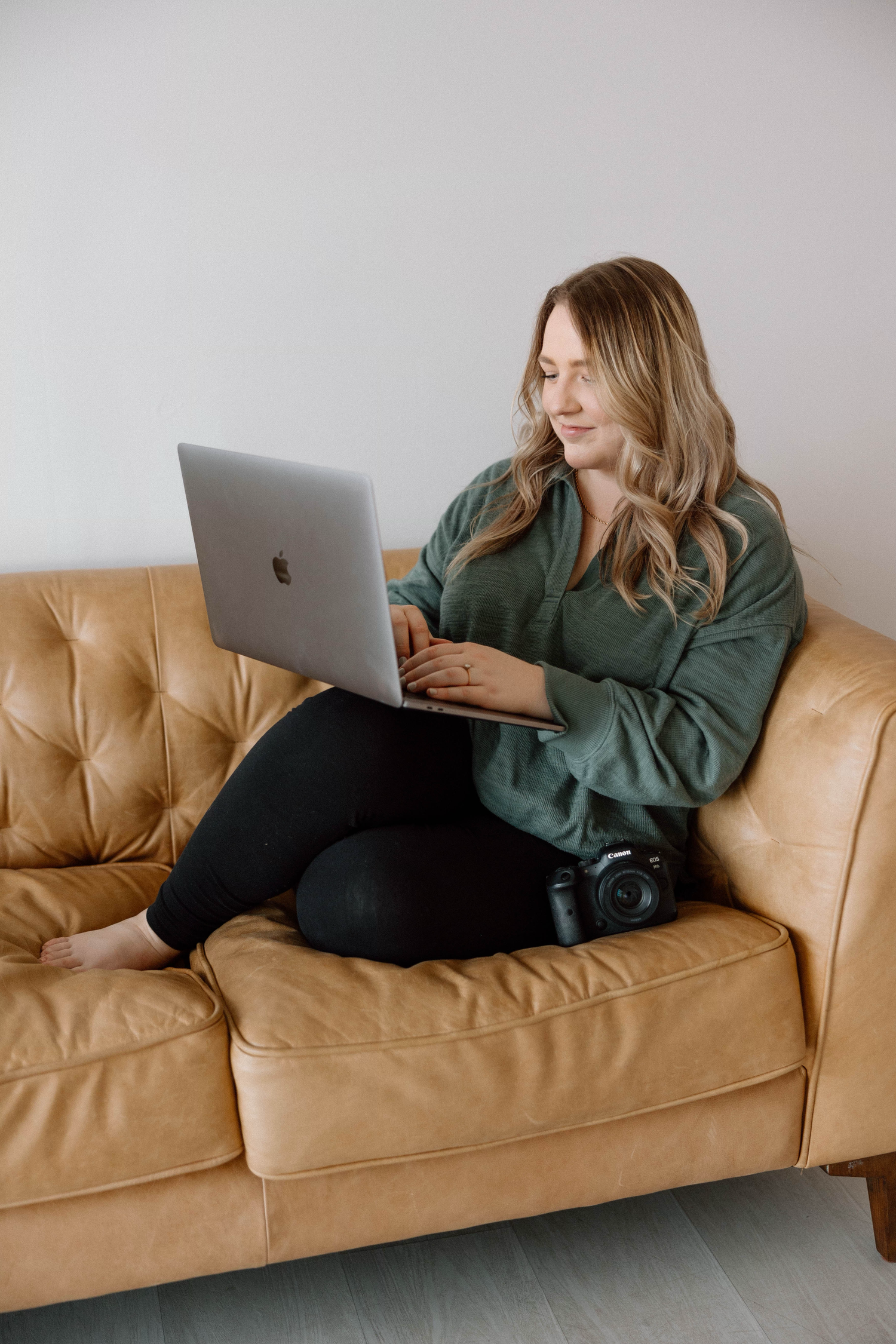Person lounging on tan leather couch with laptop and camera.