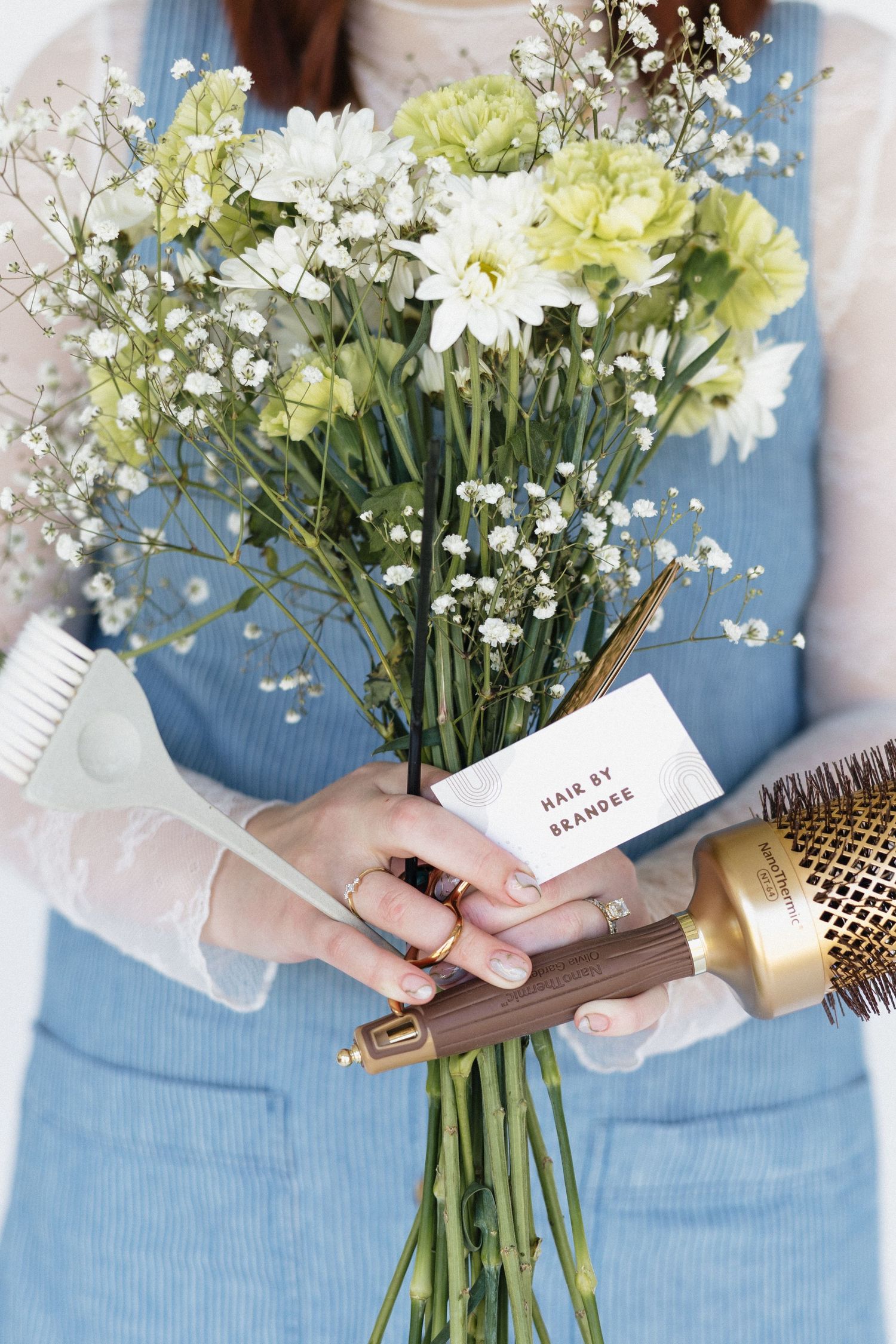 A hand holds a bouquet of white and yellow flowers with baby's breath and a small note card against a blue button-up dress.