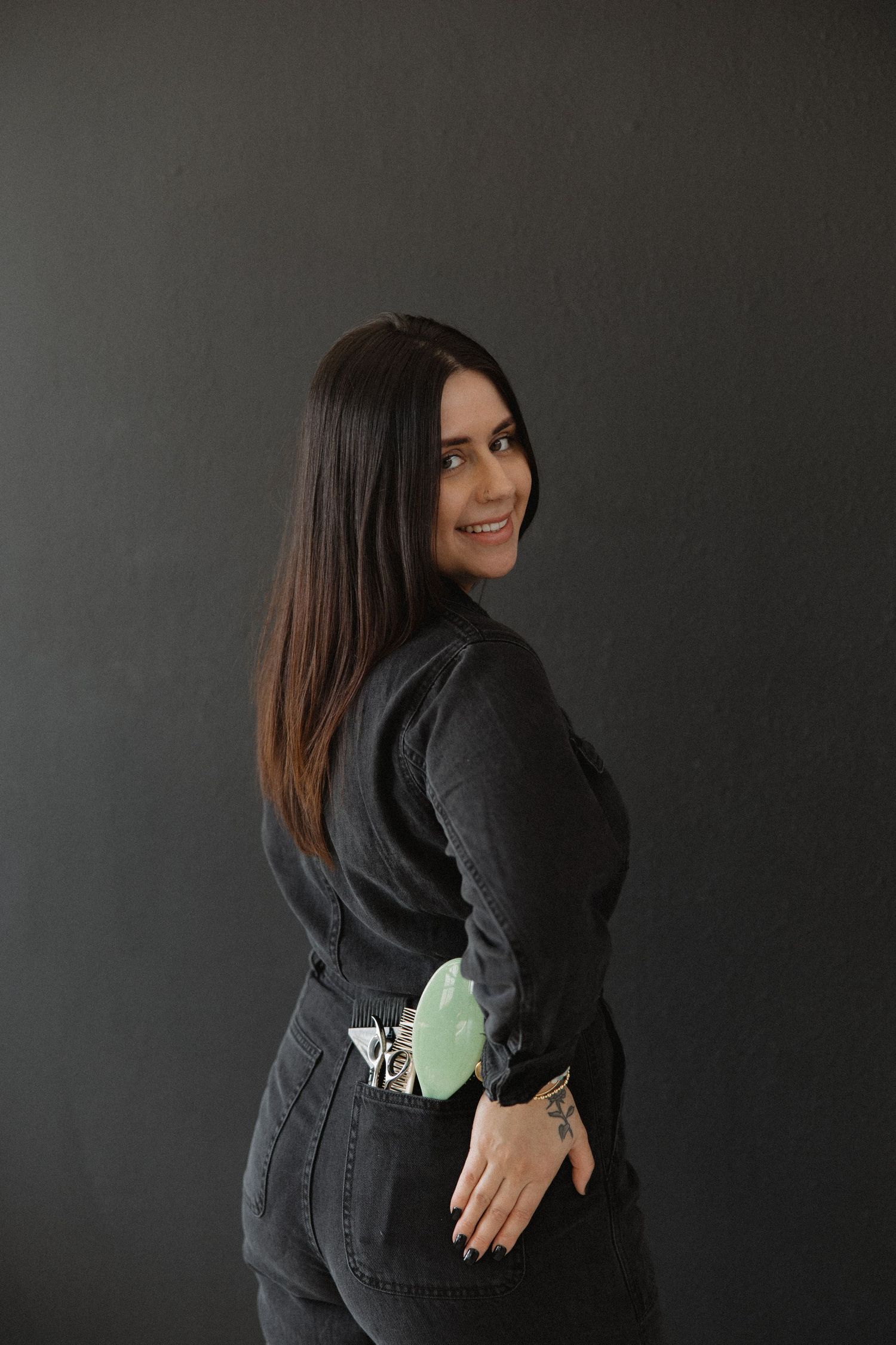 Professional wearing an all black jumpsuit poses against dark background with styling tools.