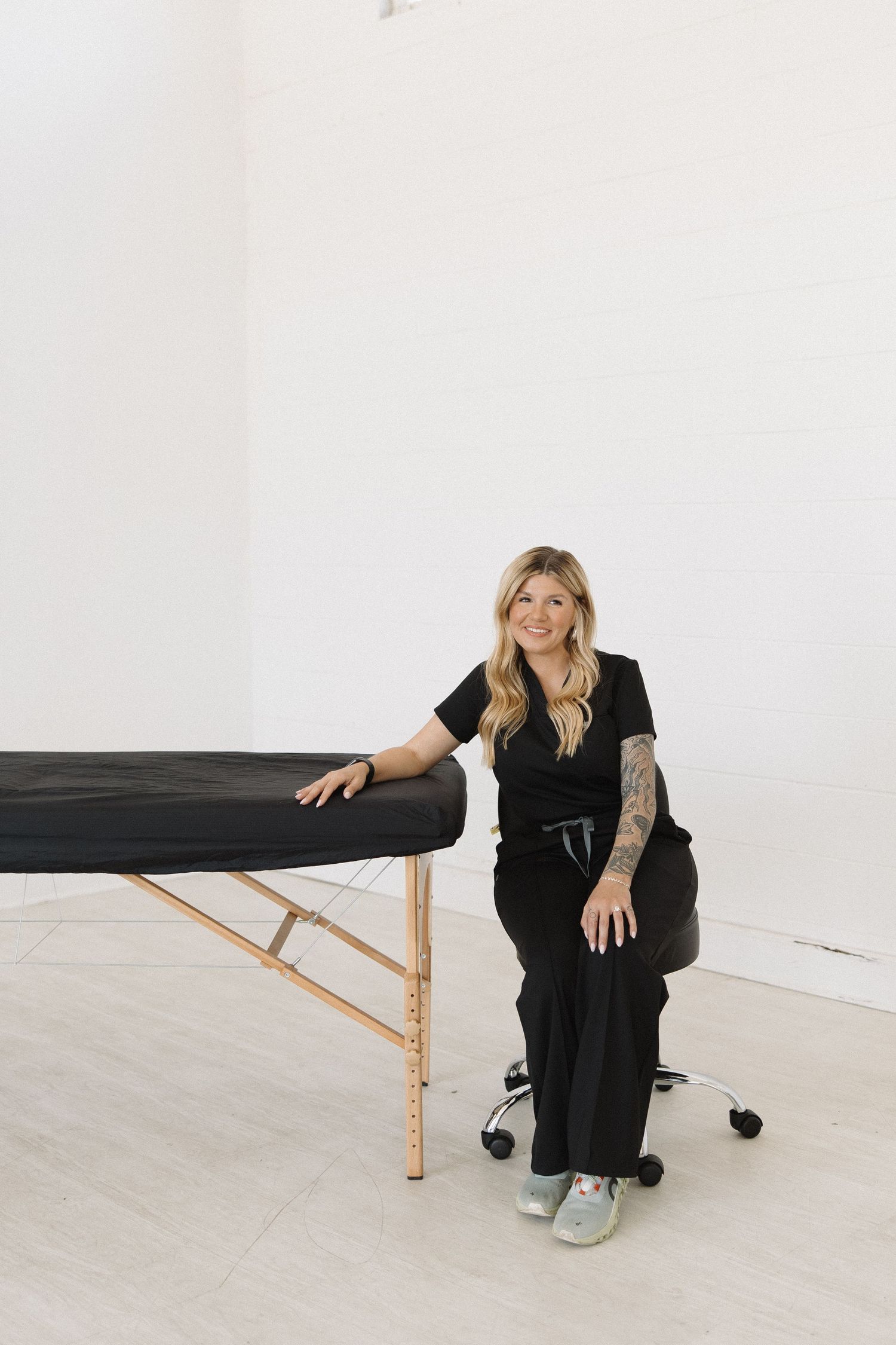 Massage therapist in black uniform sitting next to treatment table in minimalist white room.