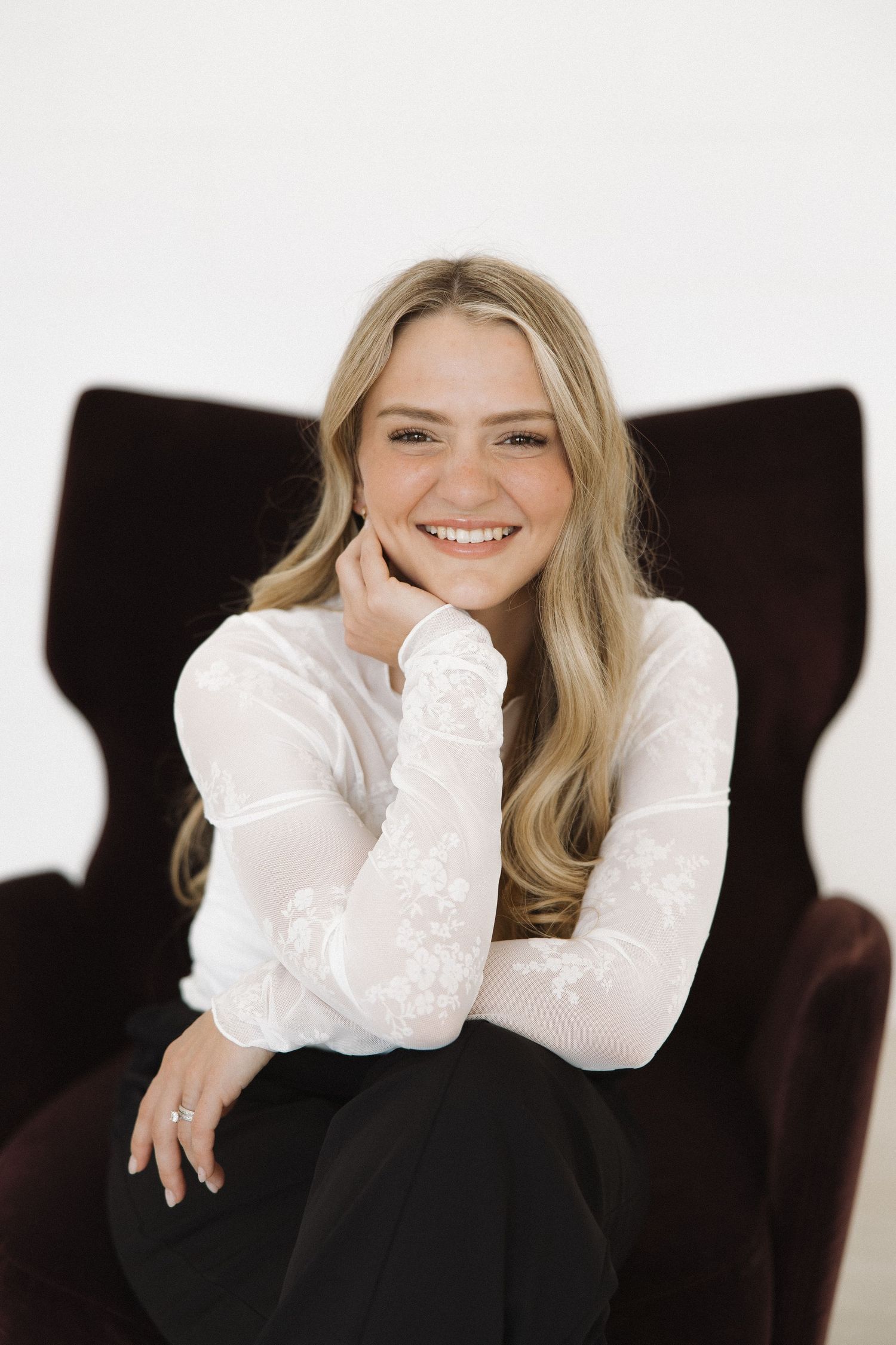 Relaxed portrait in long sleeve white top and black pants lounging on dark velvet chair.