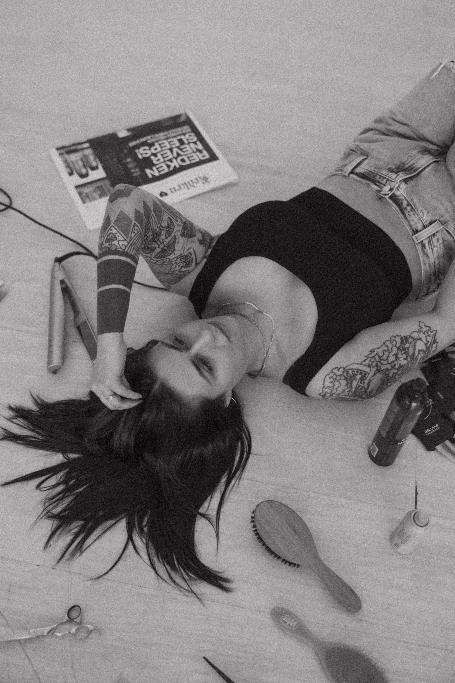 Monochrome image of tattooed person lying amid scattered hair styling tools and products.