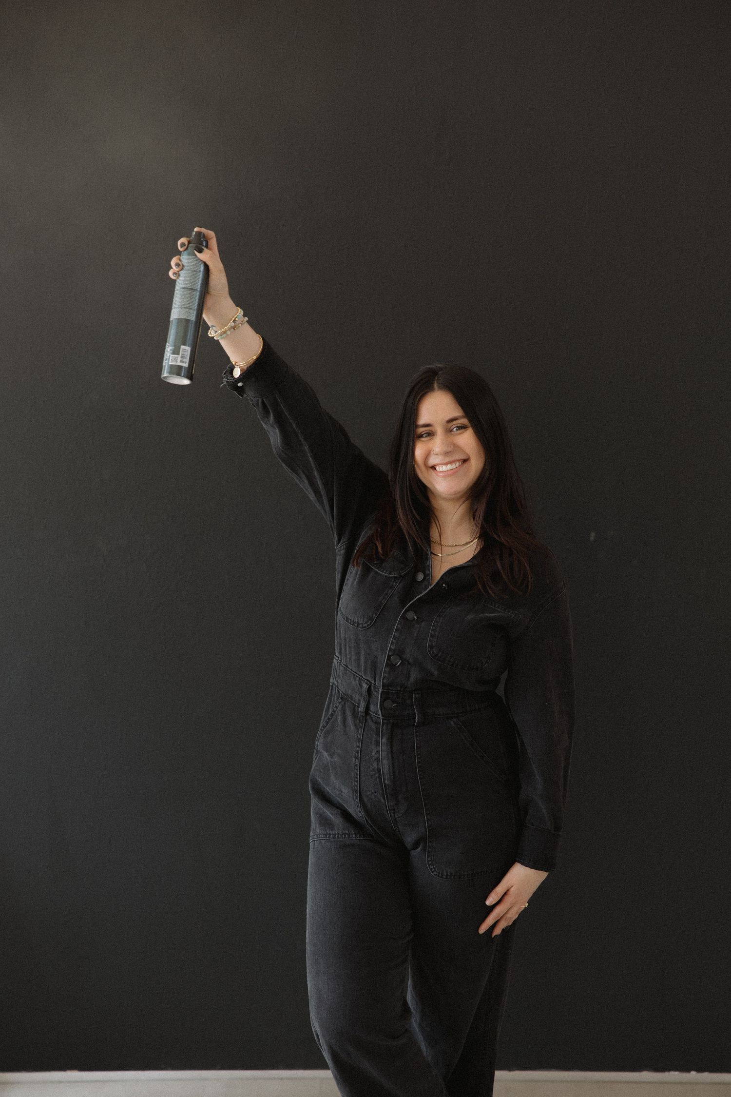 Hair professional poses with hairspray bottle while wearing black jumpsuit.