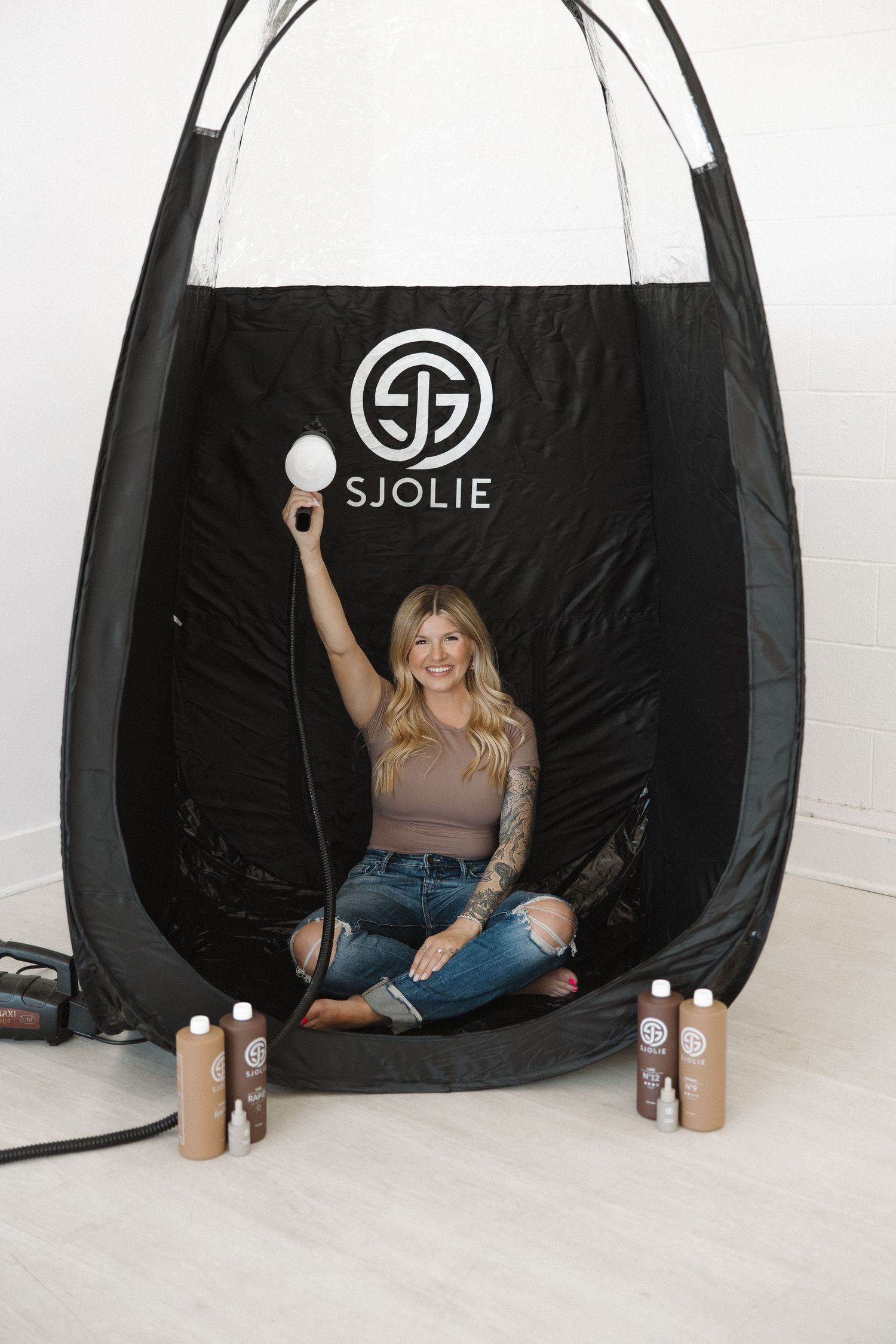 SJOLIE branded spray tan tent setup with tanning products displayed on floor.