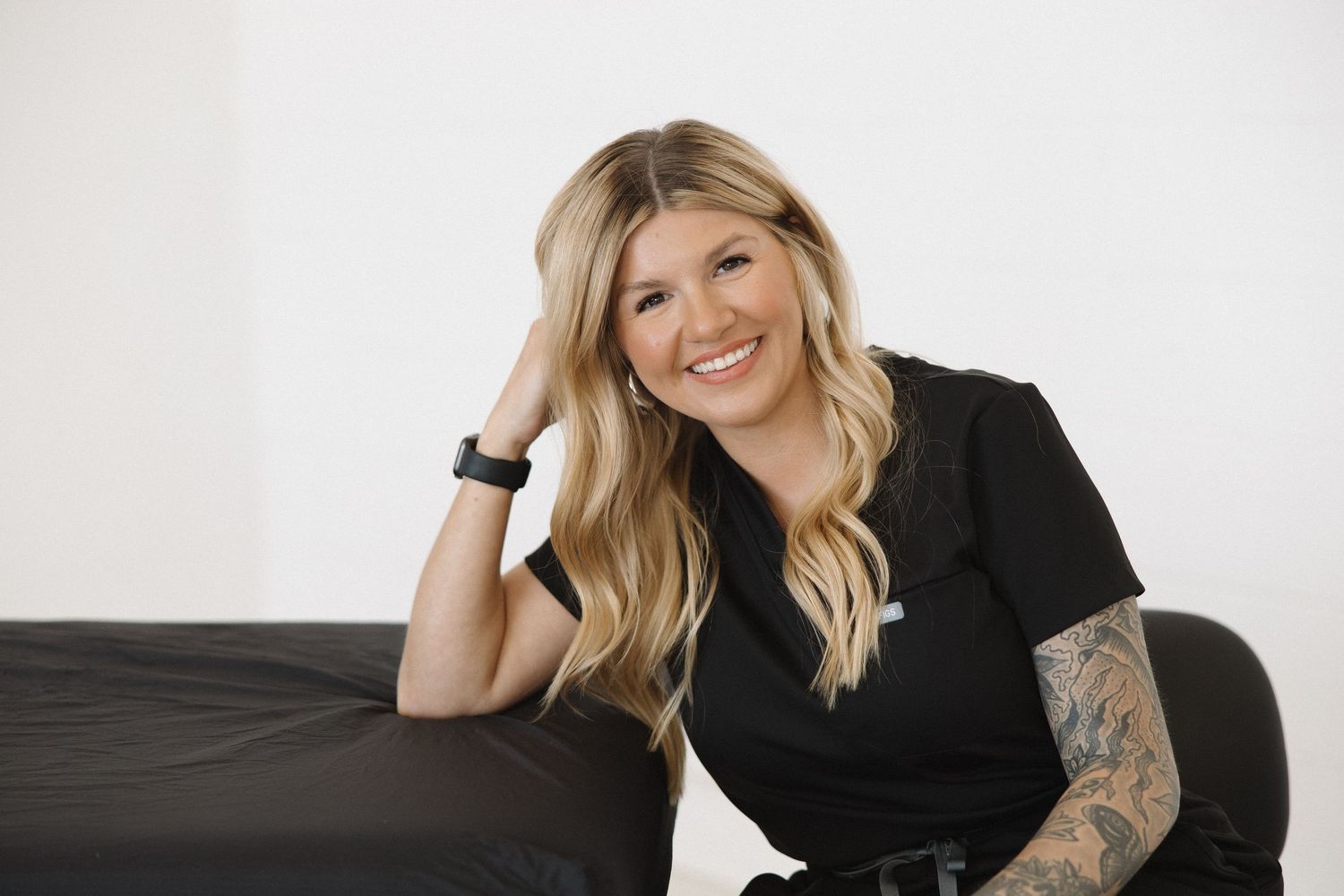 Healthcare worker in black attire sitting relaxed by massage table in bright studio setting.