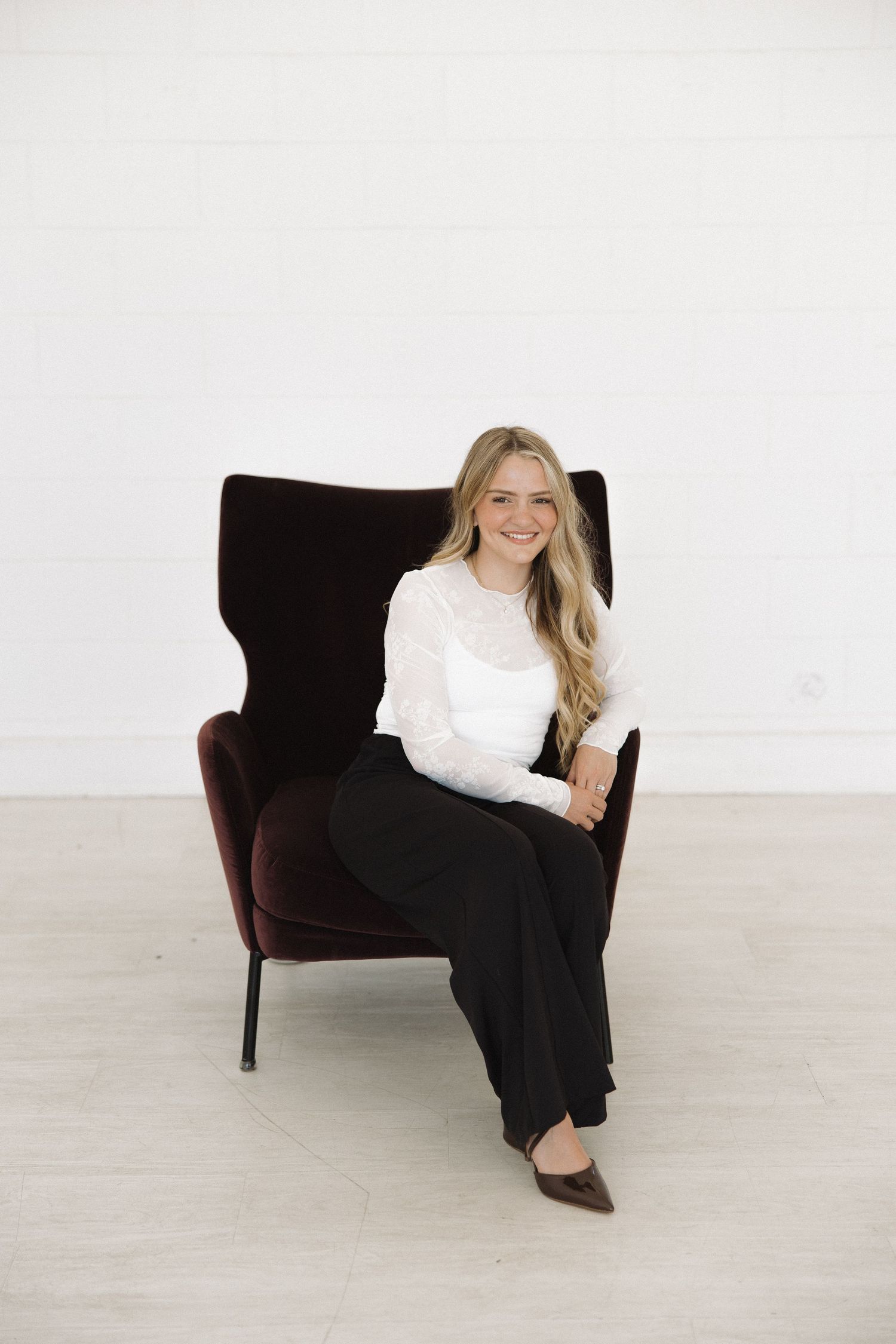 Professional portrait in classic black and white outfit seated casually on dark velvet armchair against white backdrop.