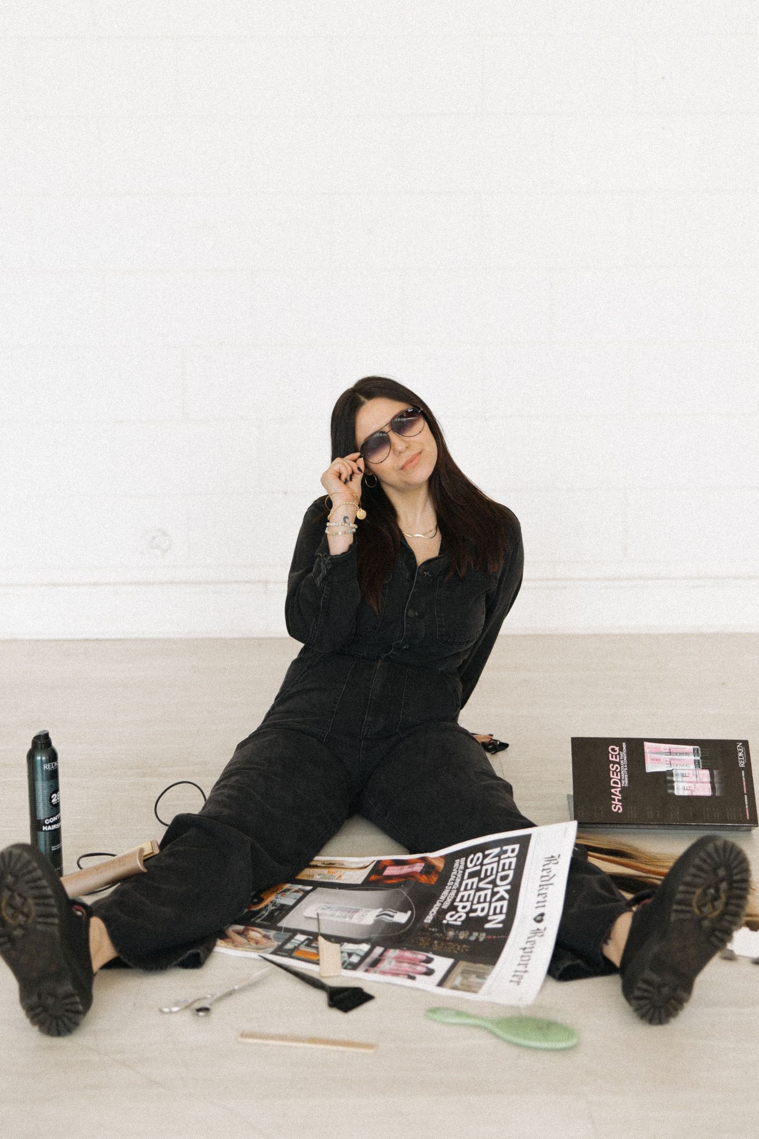 Person with brown hair in black outfit posing with magazines and styling tools on floor.