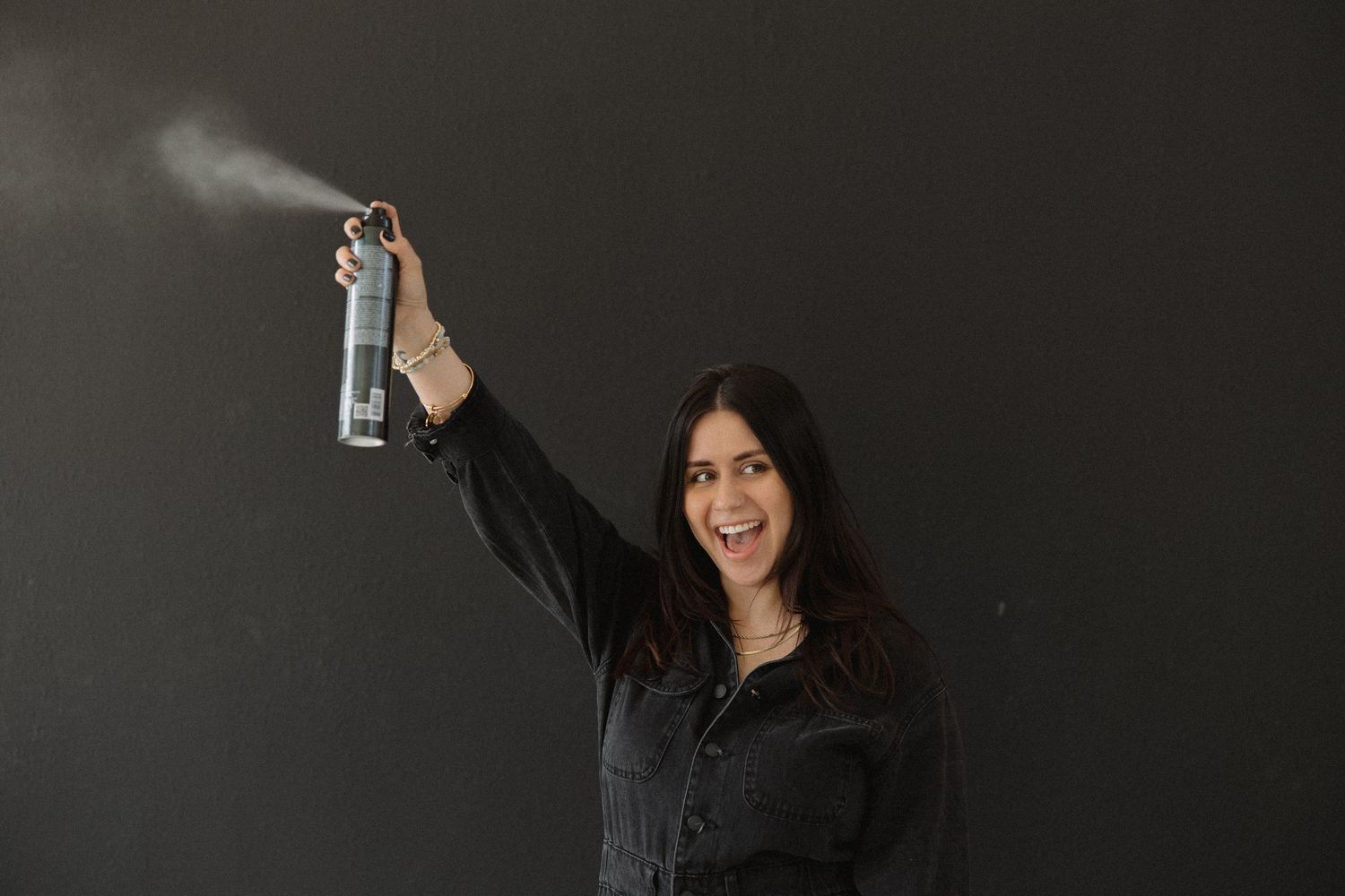 Hairstylist in black uniform holds hairspray bottle against dark wall.