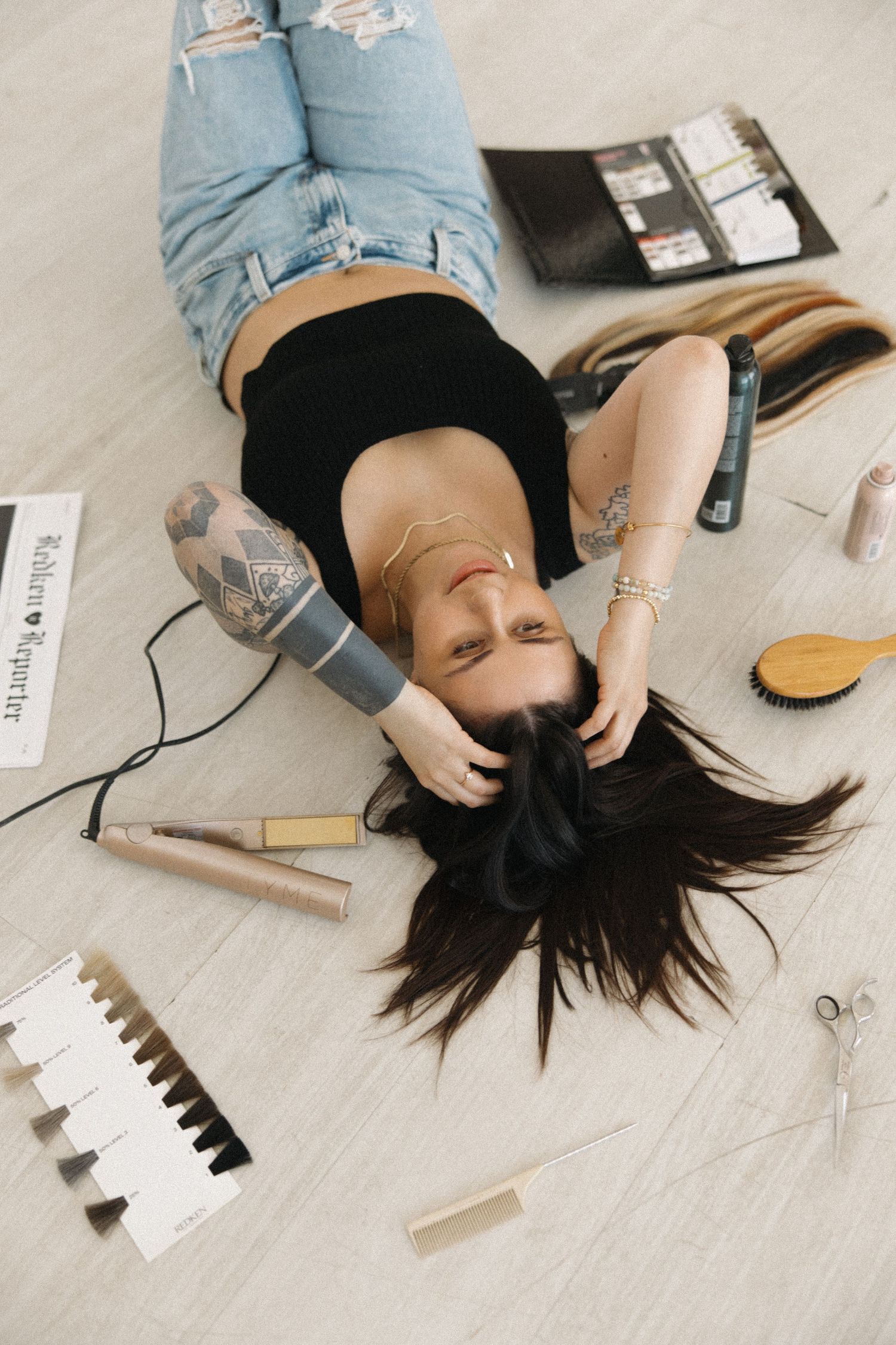 Hairstylist in denim and black lying on floor with beauty products.