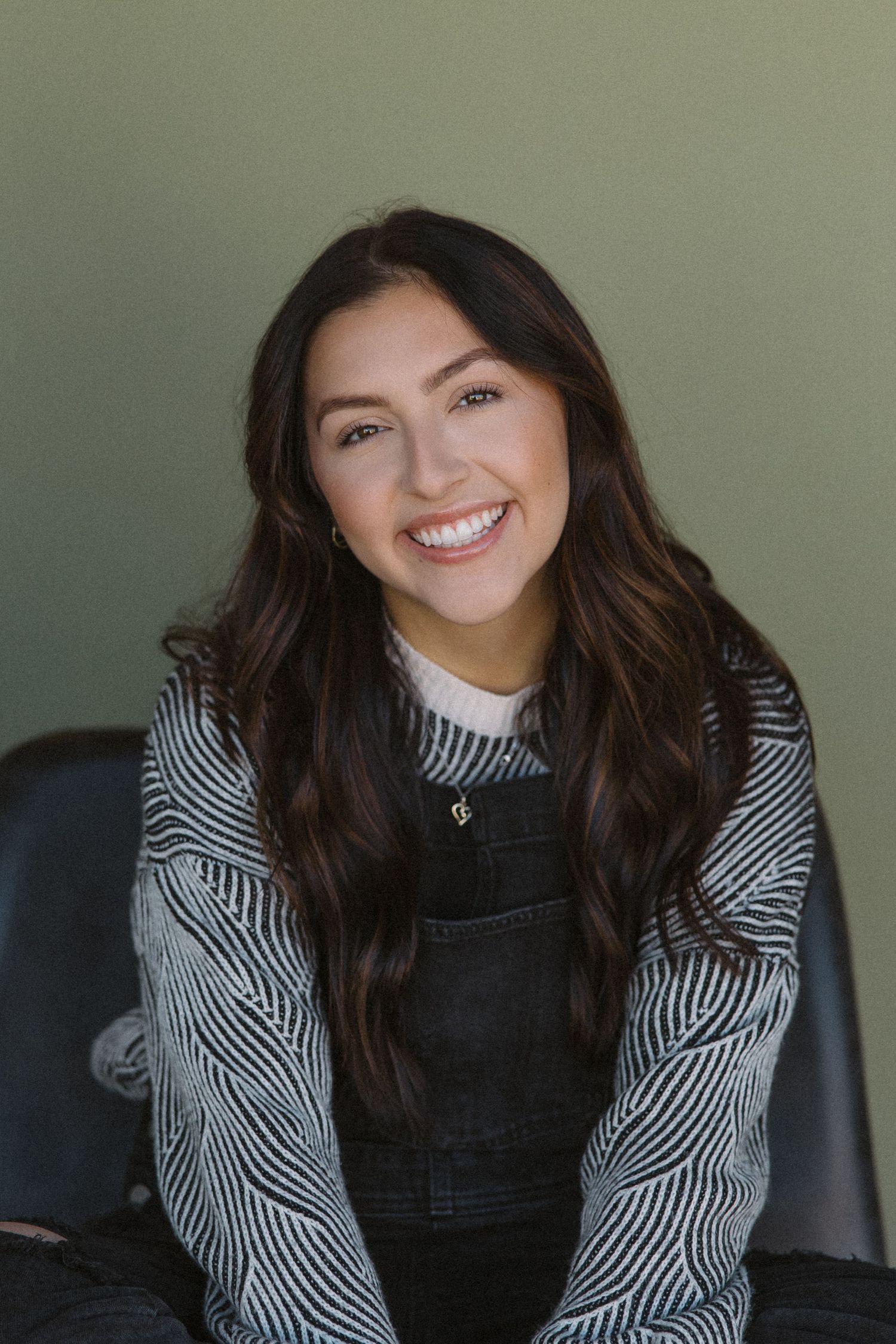 Brunette girl portrait against sage green wall wearing striped shirt and overalls.