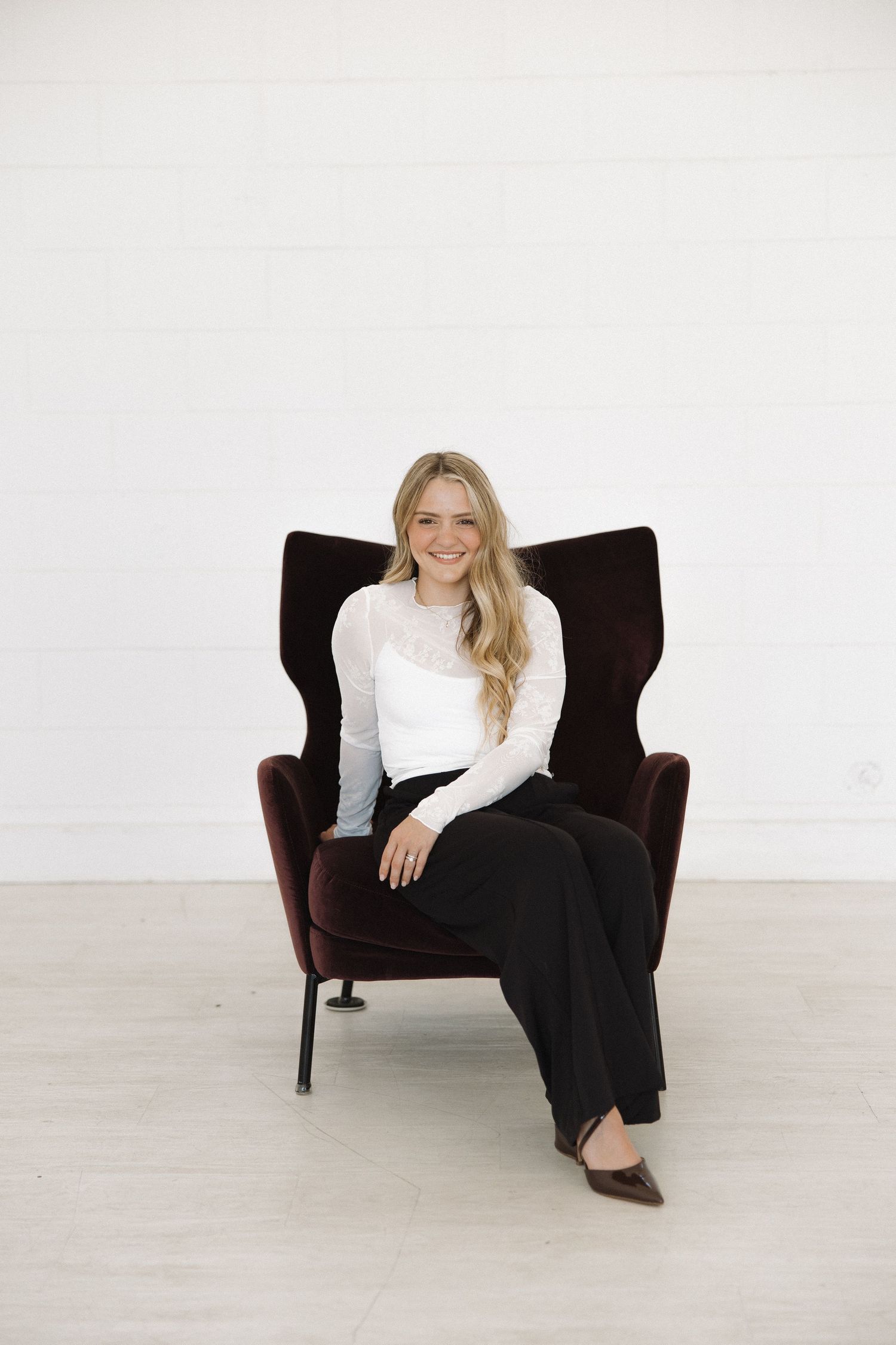 Casual lifestyle photo on dark brown armchair wearing white top and black pants in bright studio setting.