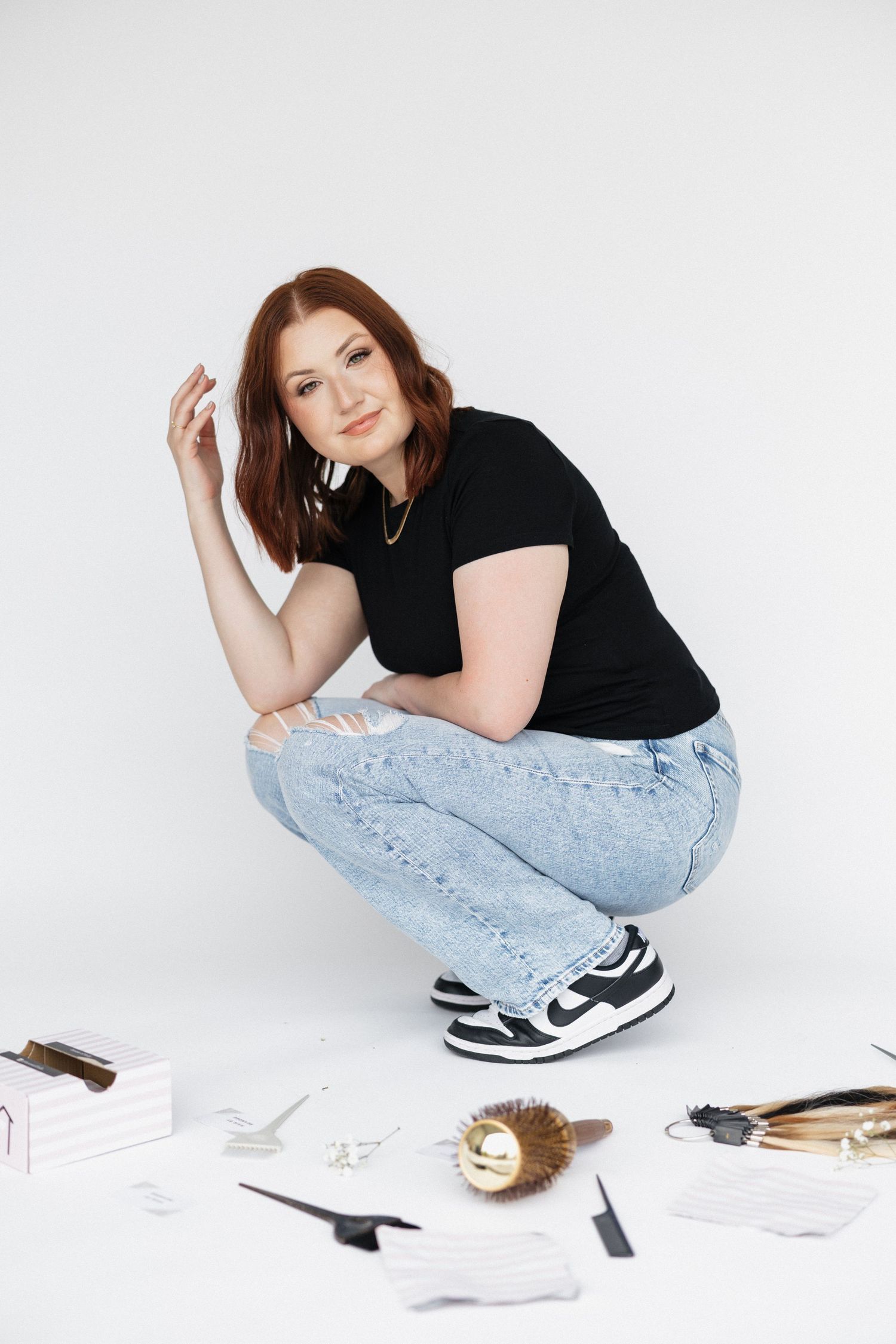 Person in black shirt and light jeans sitting with styling tools on white background.