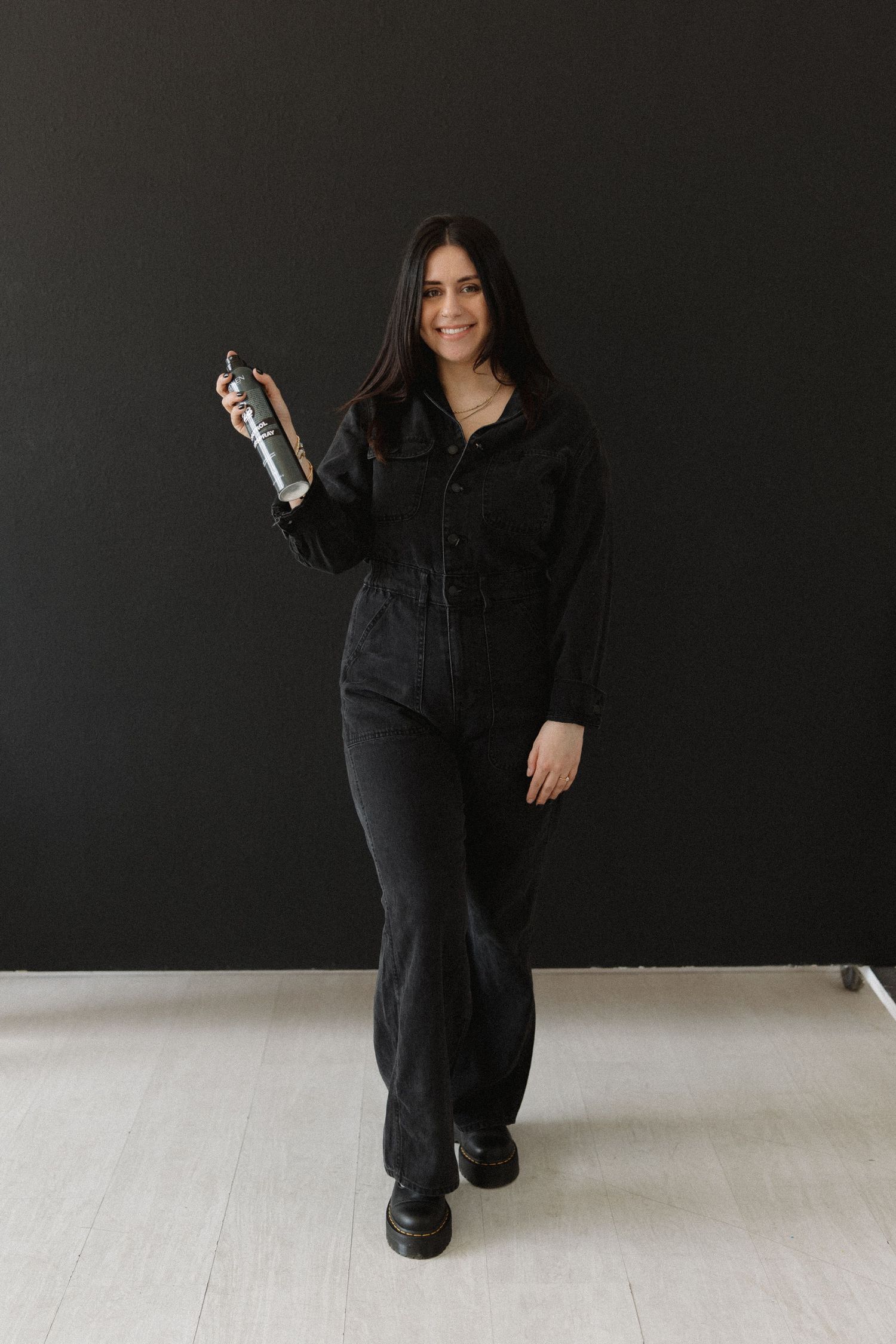 Stylist in black uniform holds hairspray bottle while standing against dark wall.