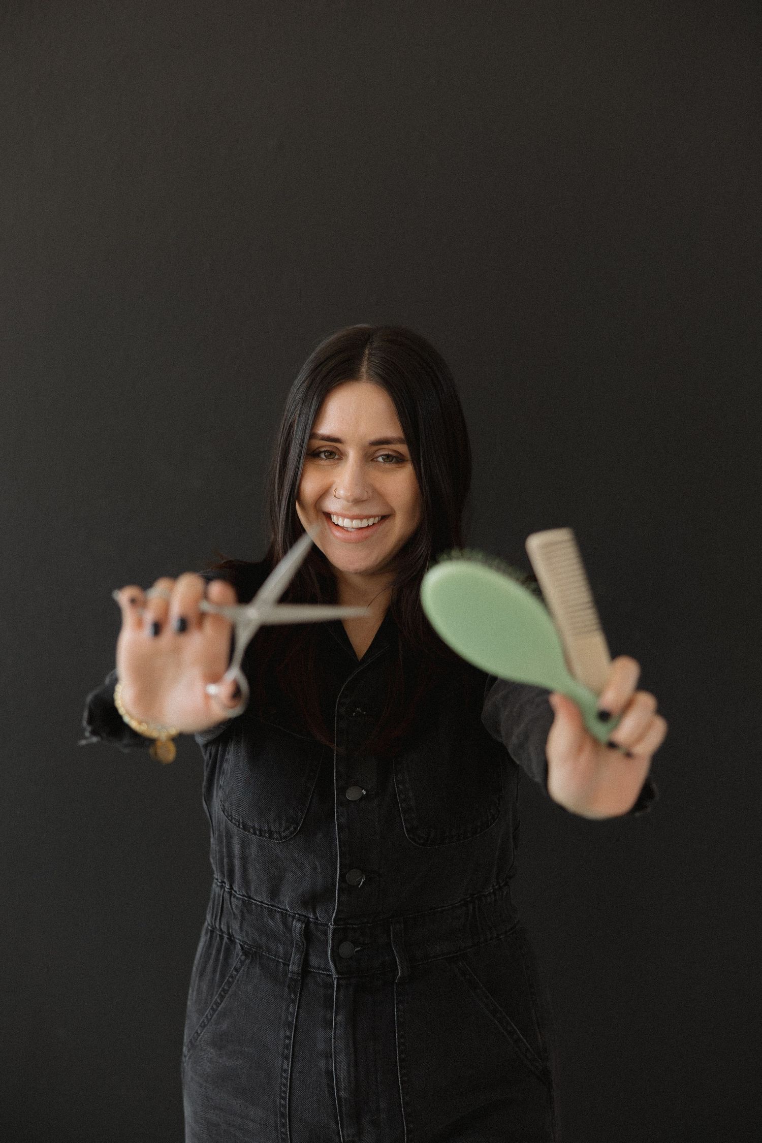 Stylist holds up hair tools while wearing black jumpsuit against dark background.