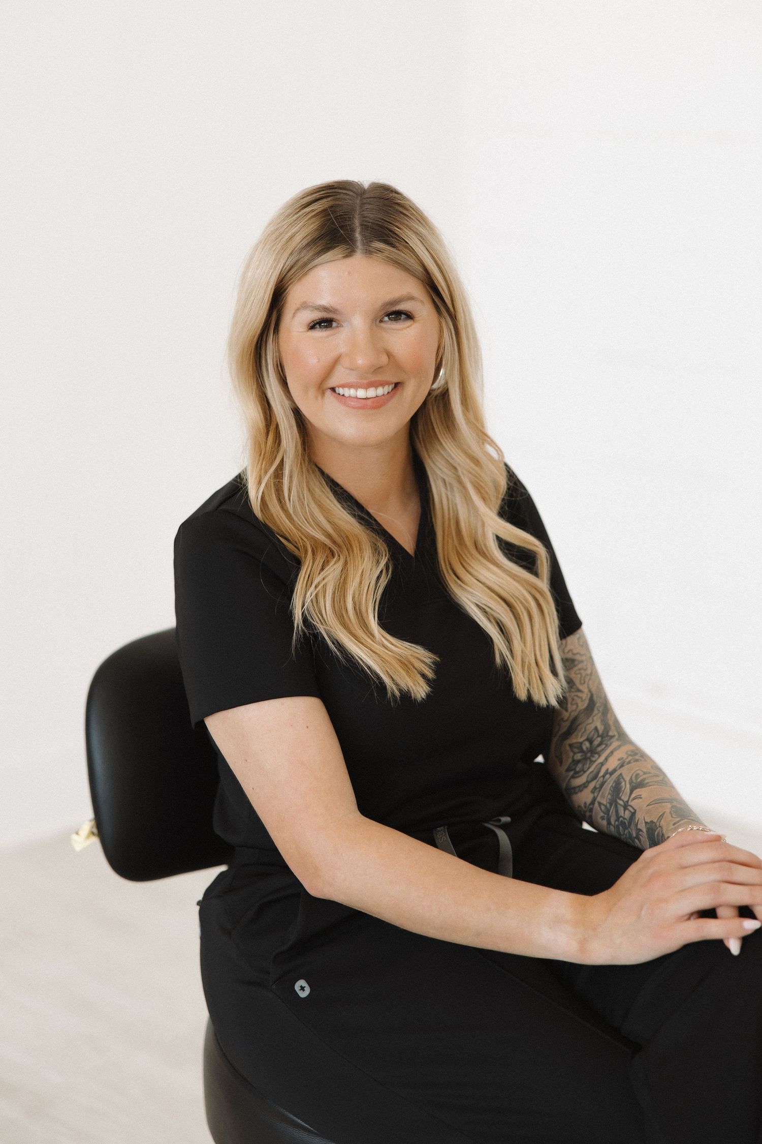 Blonde-haired individual in black outfit poses professionally in white studio setting.