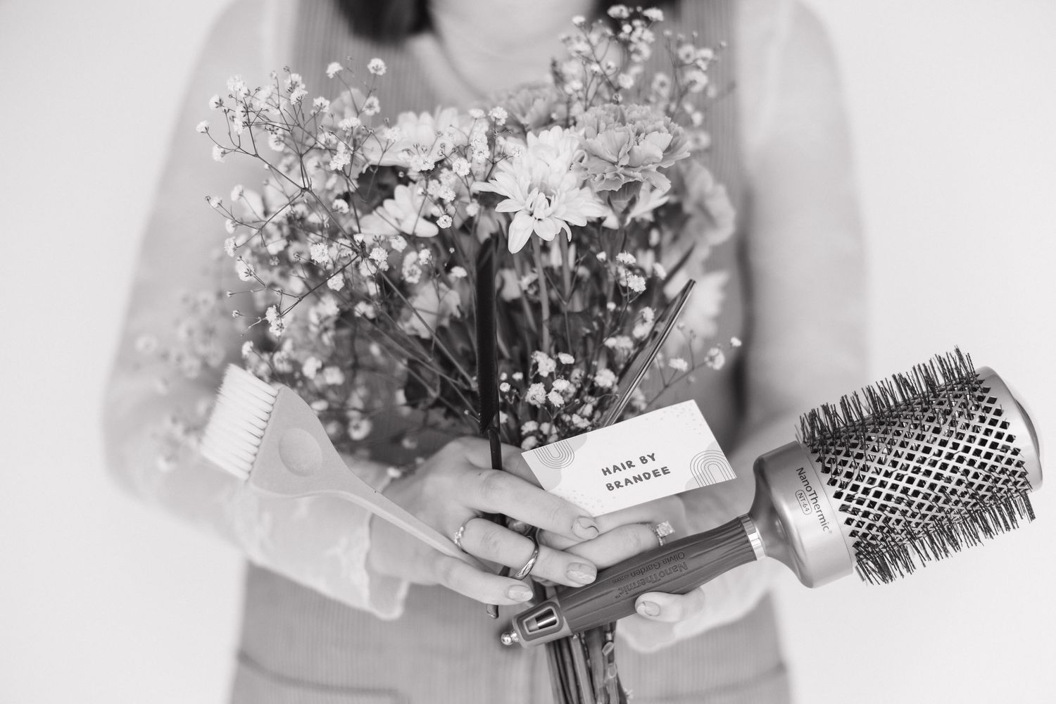 Black and white image of hands holding flowers and hair styling tools against a light background.