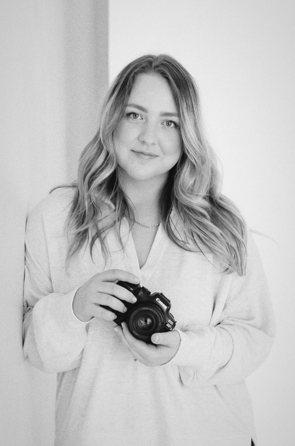 Person in white outfit holding camera against light background.