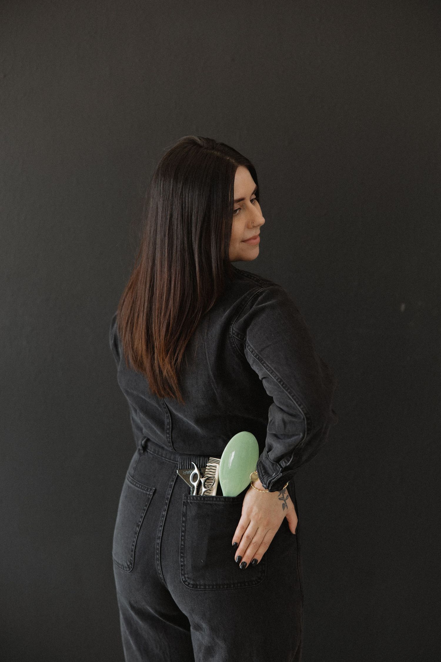 Stylist in black denim outfit shows off styling tools against dark backdrop.