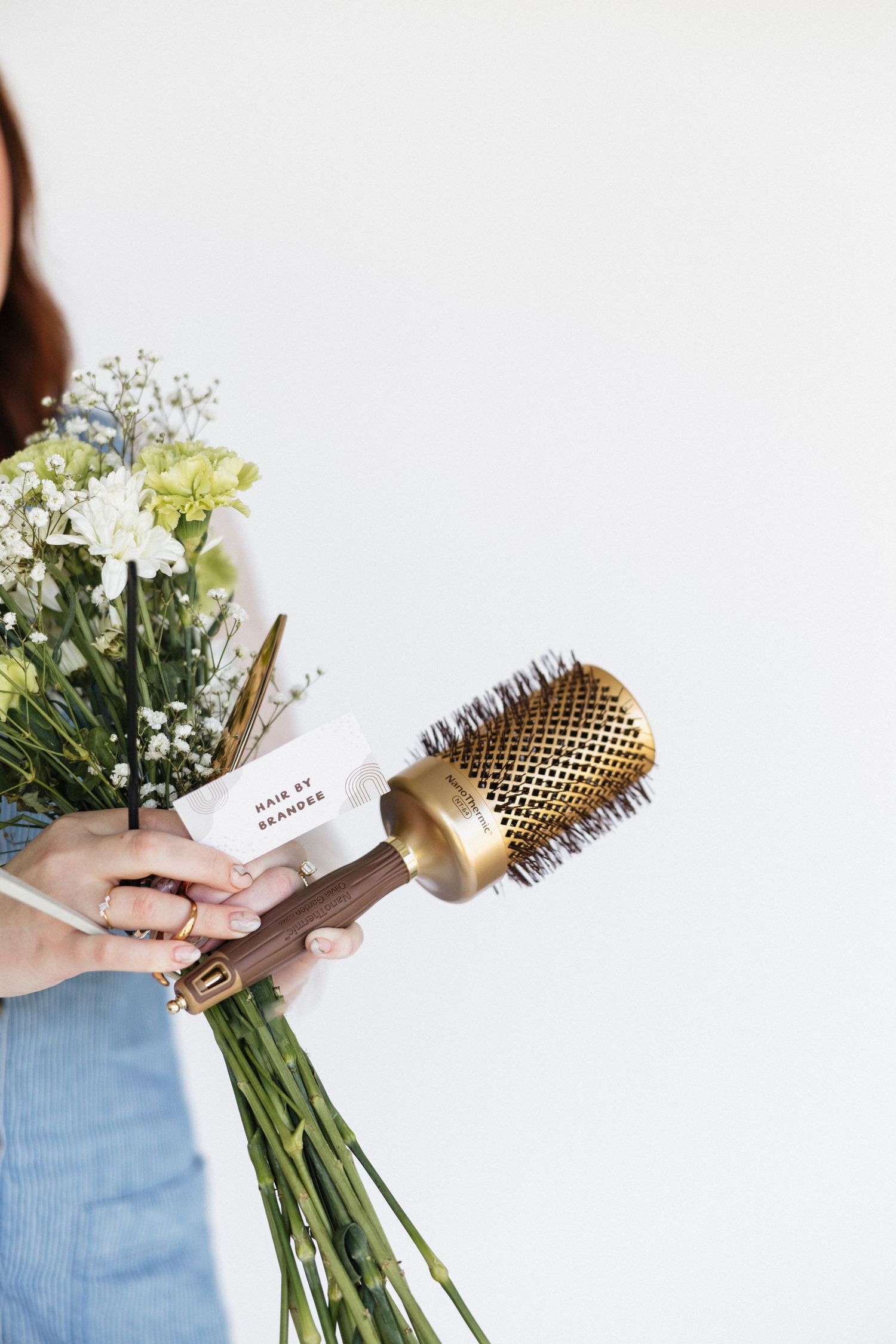 Hands holding a fresh flower bouquet and gold styling brush against white background.