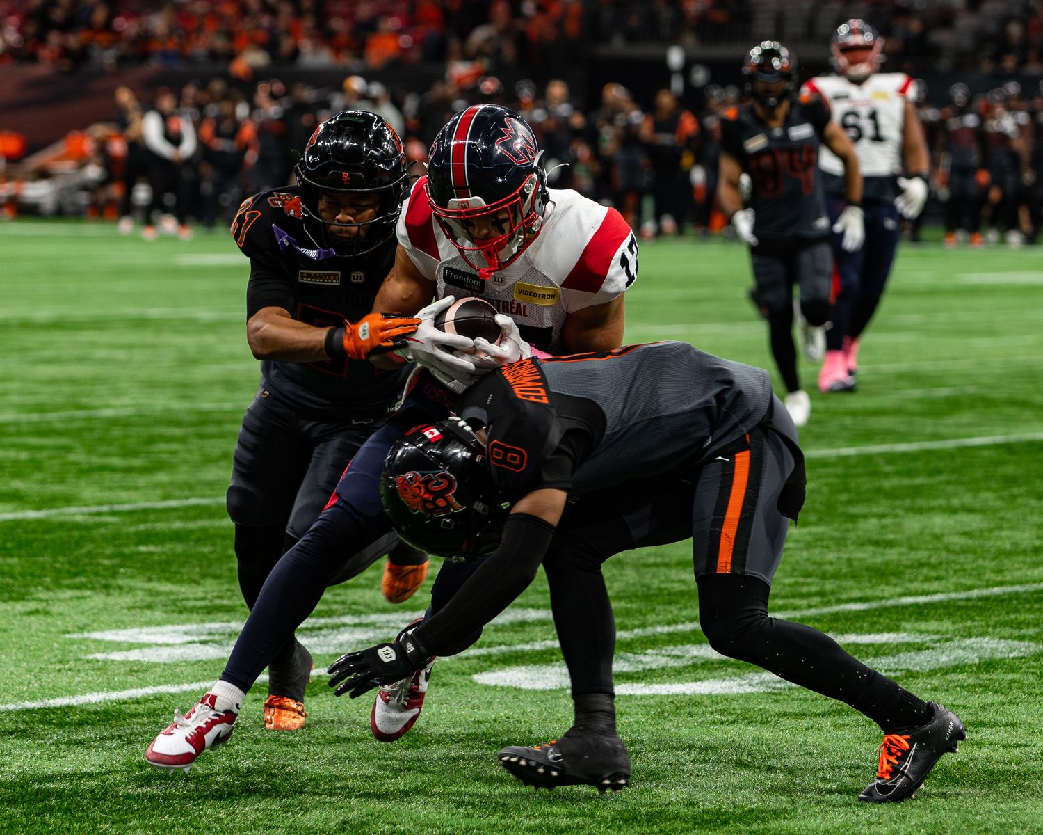 Football players in orange and black uniforms tackle a player in red and white during an indoor CFL game.